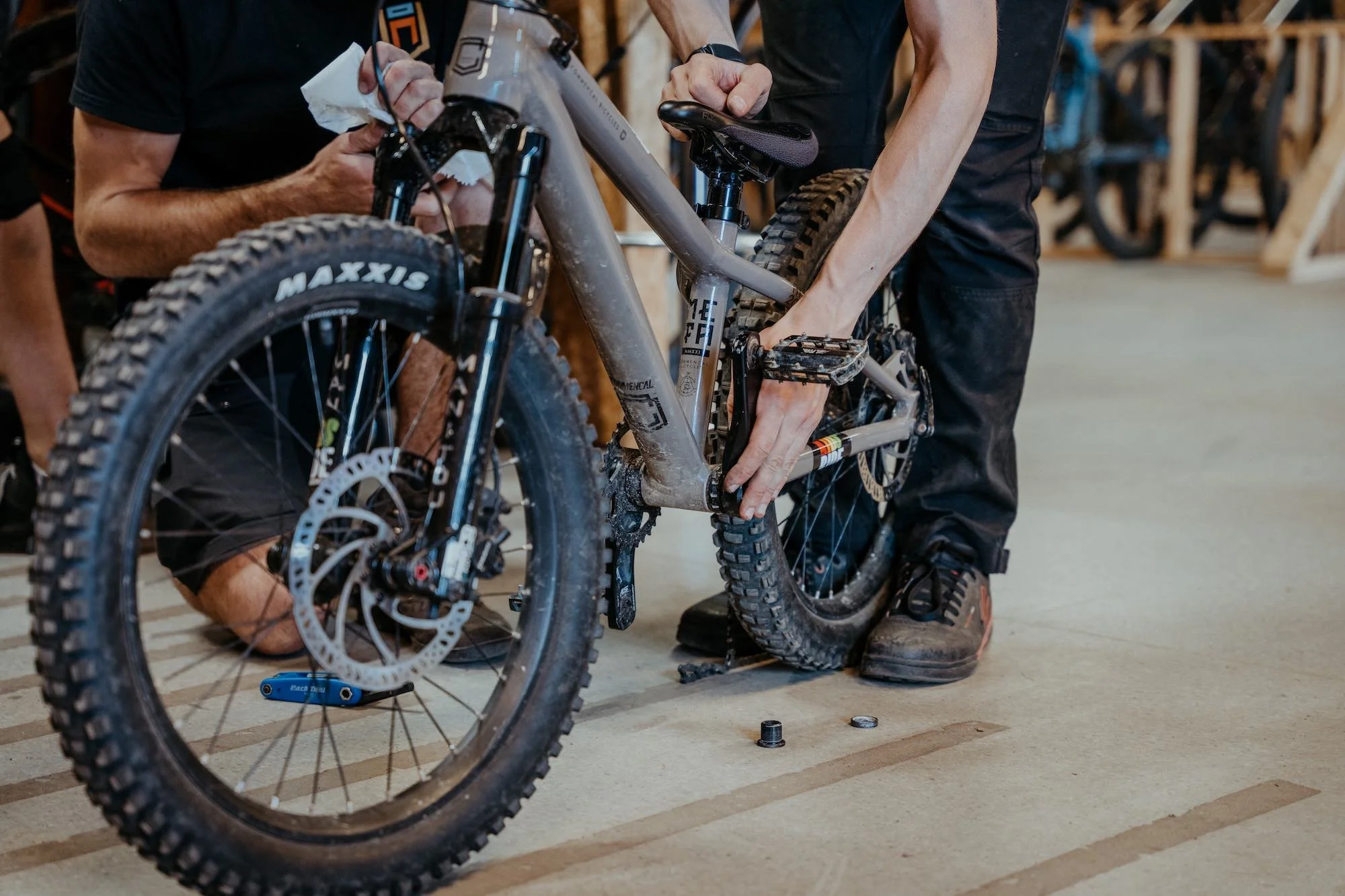 Två personer reparerar en mountainbike inomhus. En person står medan den andra justerar cykelns nedre del. Cykeln är dammig och står på ett betonggolv.