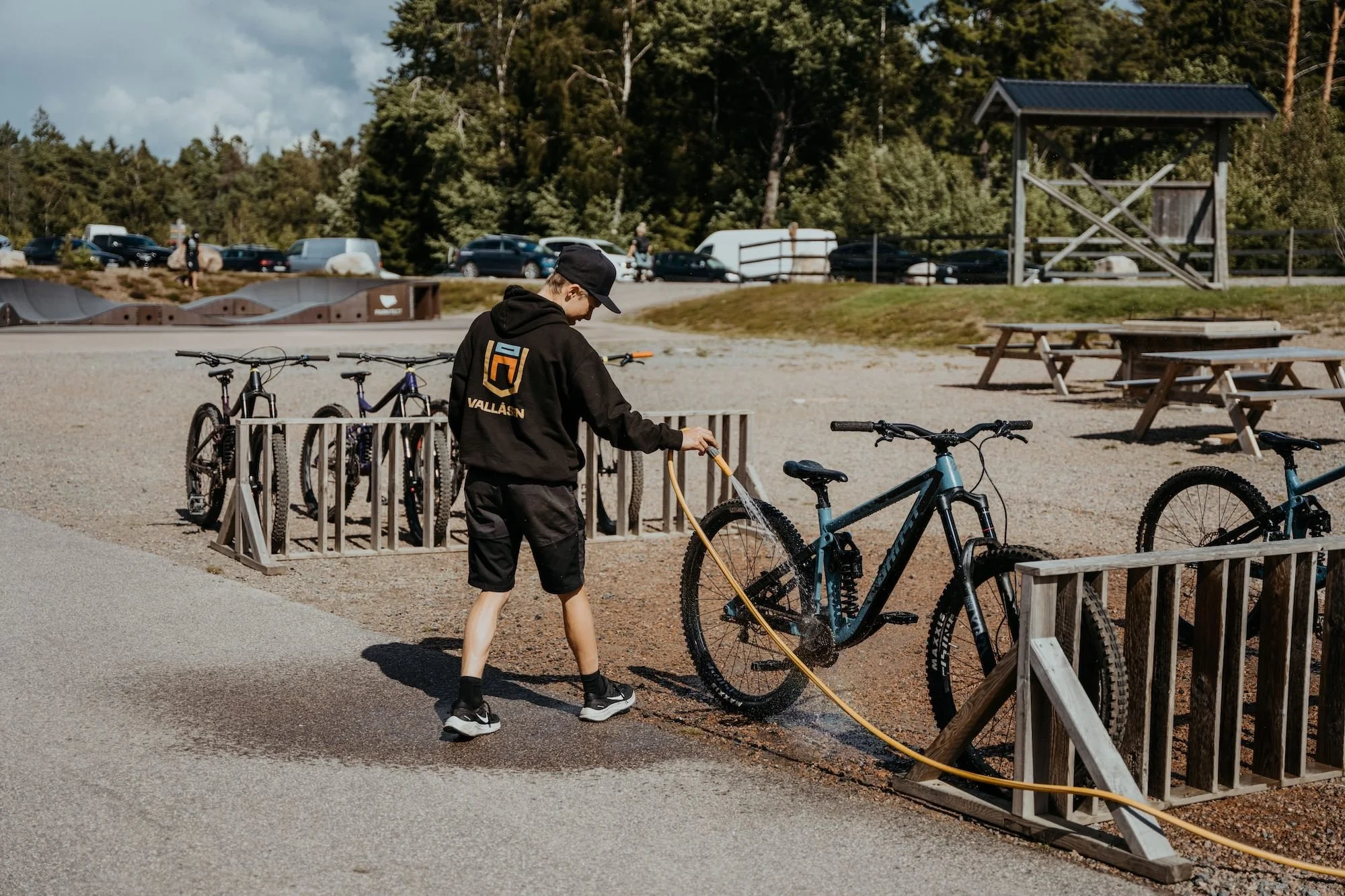 En pojke tvättar sin cykel utomhus på Vallåsen Bike Park, fler cyklar står parkerade i bakgrunden och det finns träd samt parkbänkar i omgivningen.