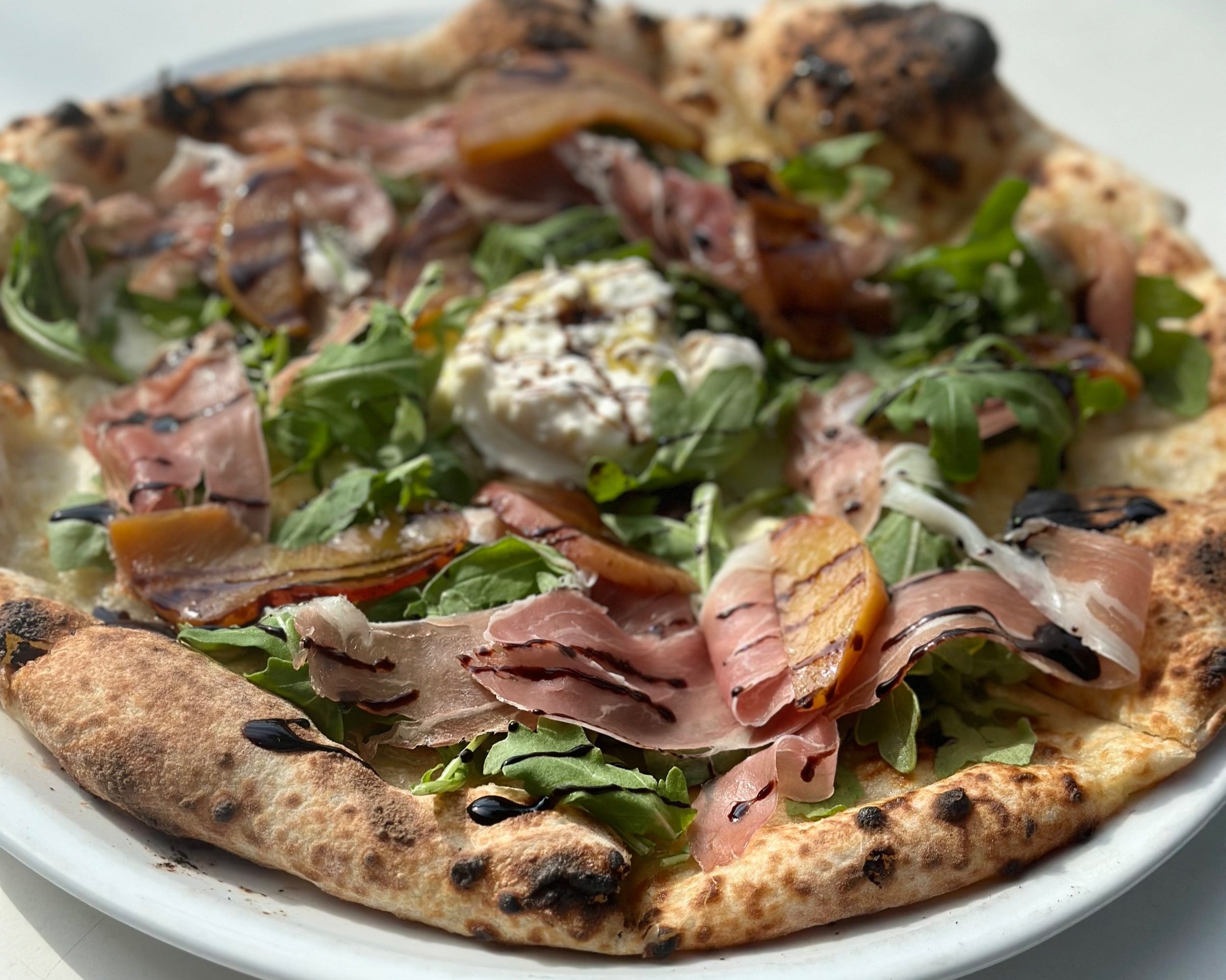 Close-up of a gourmet pizza topped with arugula, thin slices of prosciutto, grilled chicken, dollops of cheese, and a drizzle of balsamic glaze, served on a white plate.