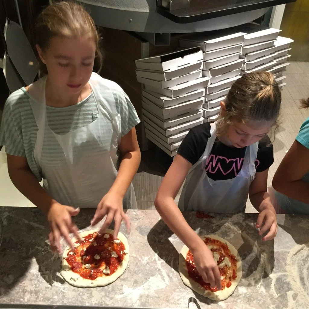 Two young girls are making mini pizzas with tomato sauce and cheese on a marble countertop in a kitchen or pizzeria. There are stacked boxes in the background.