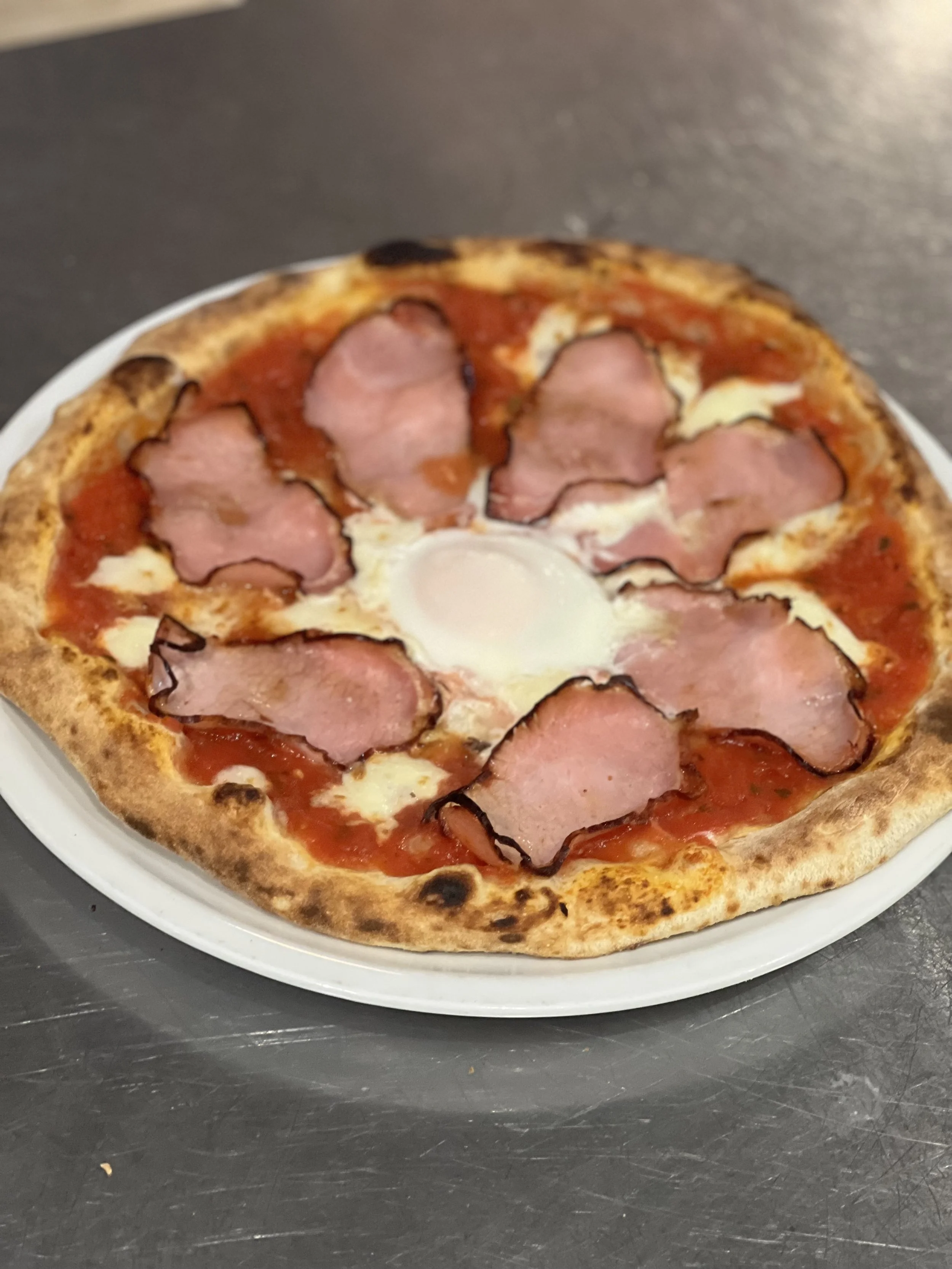 Pizza with ham slices and an egg in the center on a white plate