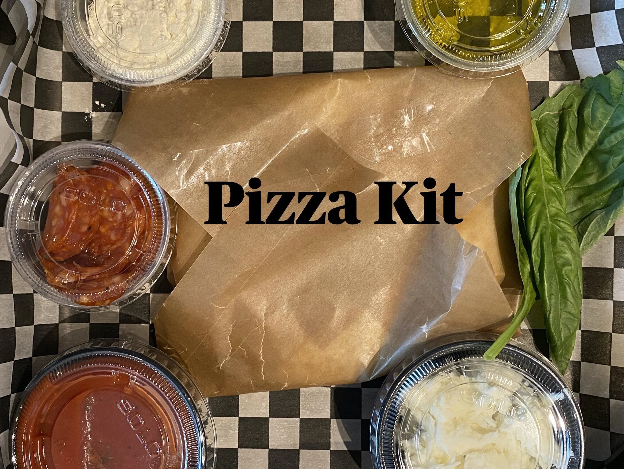 A pizza kit with ingredients including basil leaves and various toppings in plastic containers, placed on a checkered tablecloth with a brown paper bag in the center labeled 'Pizza Kit'.