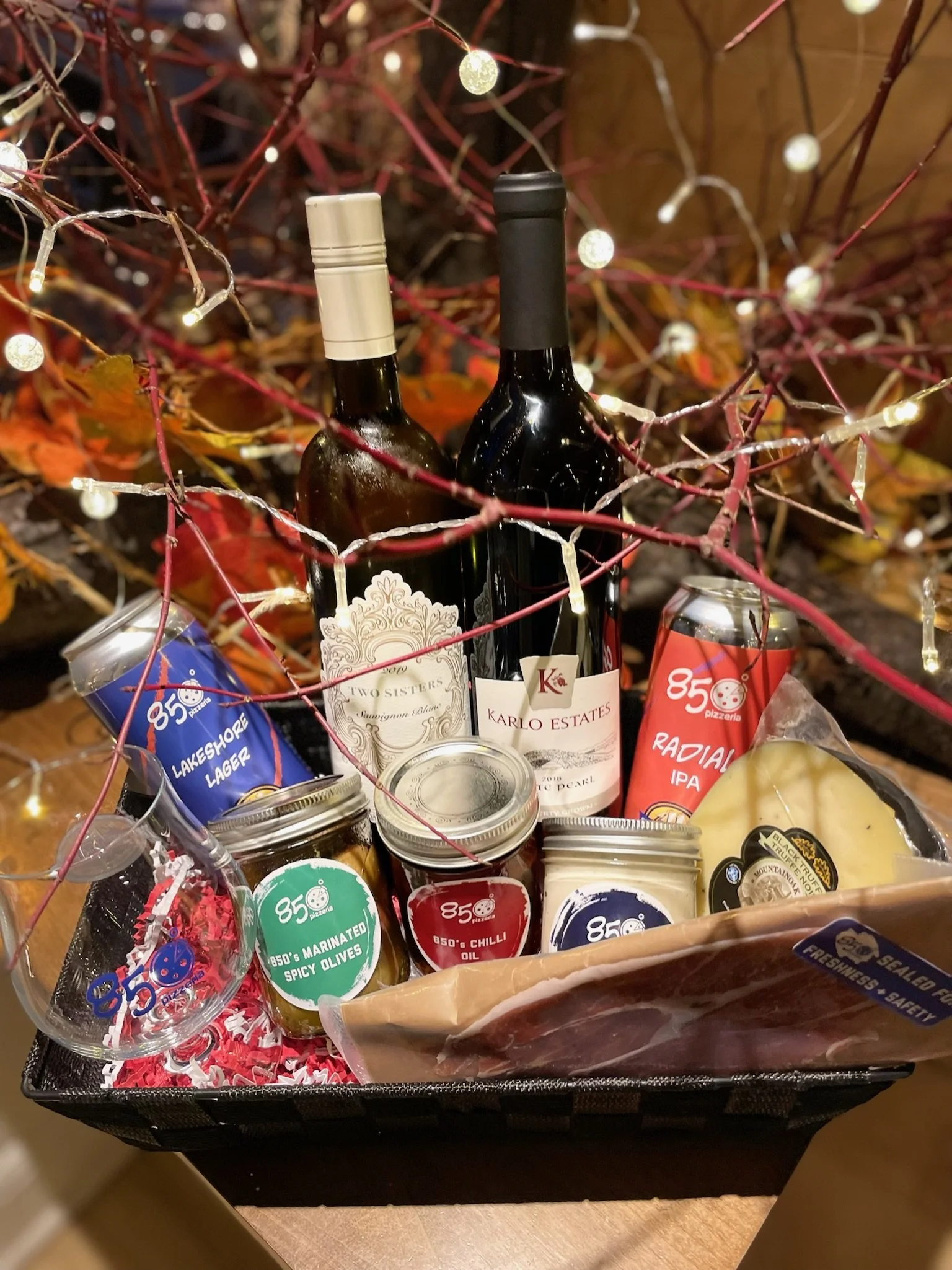 A gift basket with two bottles of wine, snacks, canned beverages, and other food items, decorated with string lights and red branches with leaves.