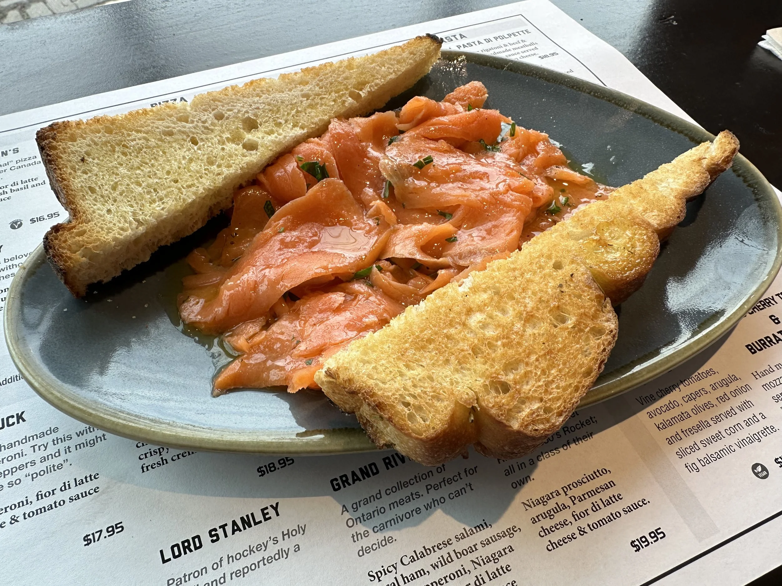 Piece of smoked salmon, two toasted bread slices, and chopped herbs on a dark ceramic plate on a restaurant menu