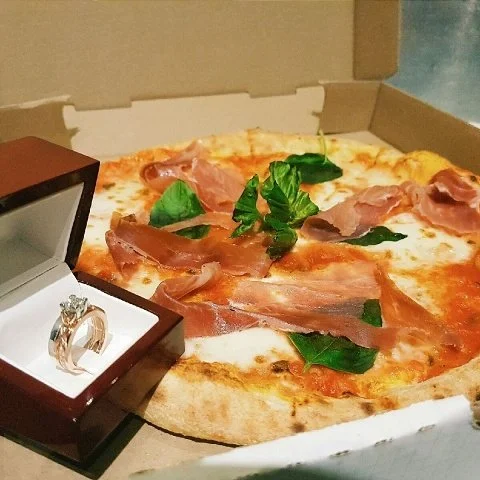 A pizza with ham and leafy greens in an open pizza box, with a ring in a jewelry box placed on the pizza.