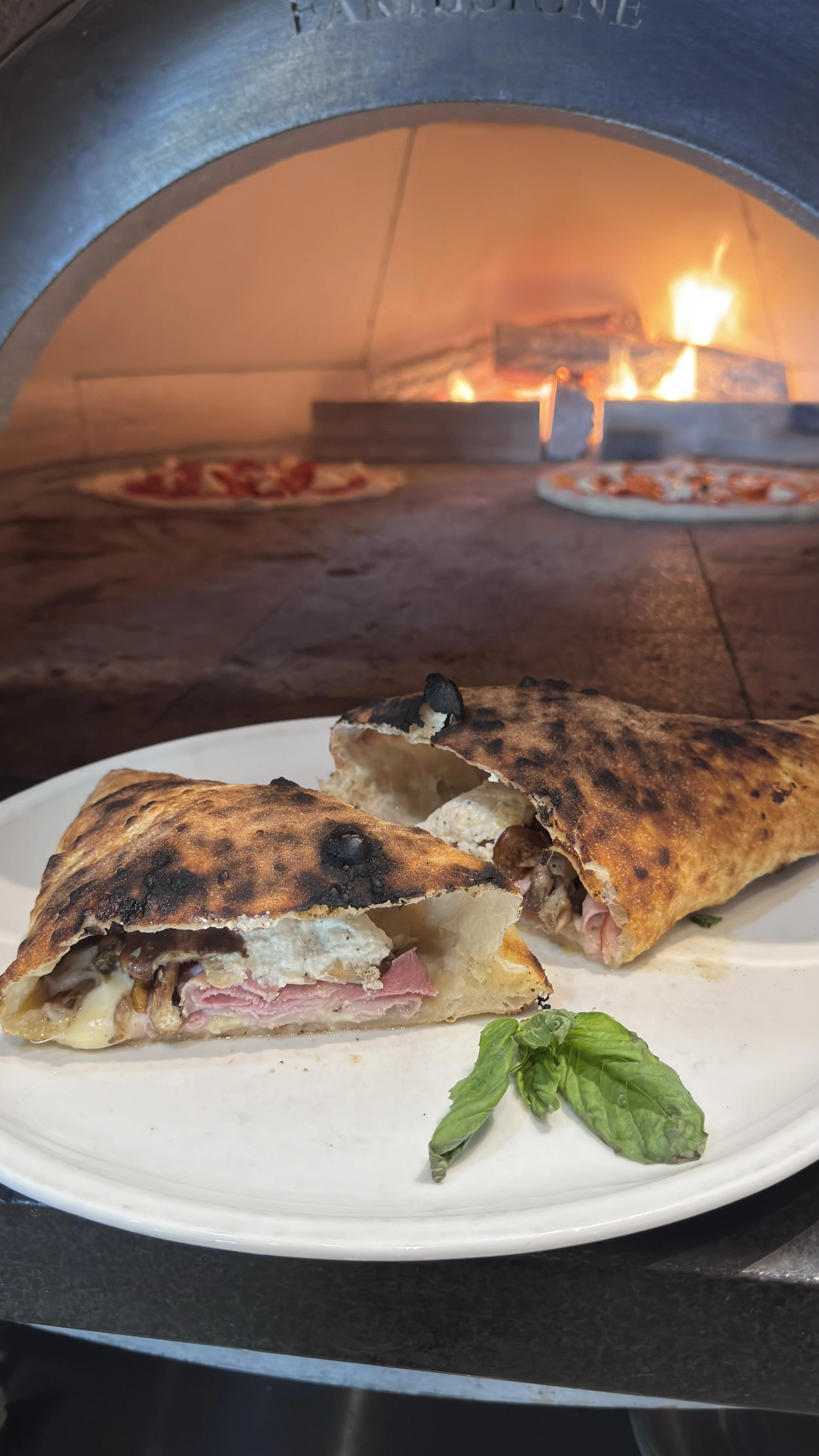 Calzone on a white plate with basil, with a wood-fired pizza oven in the background.