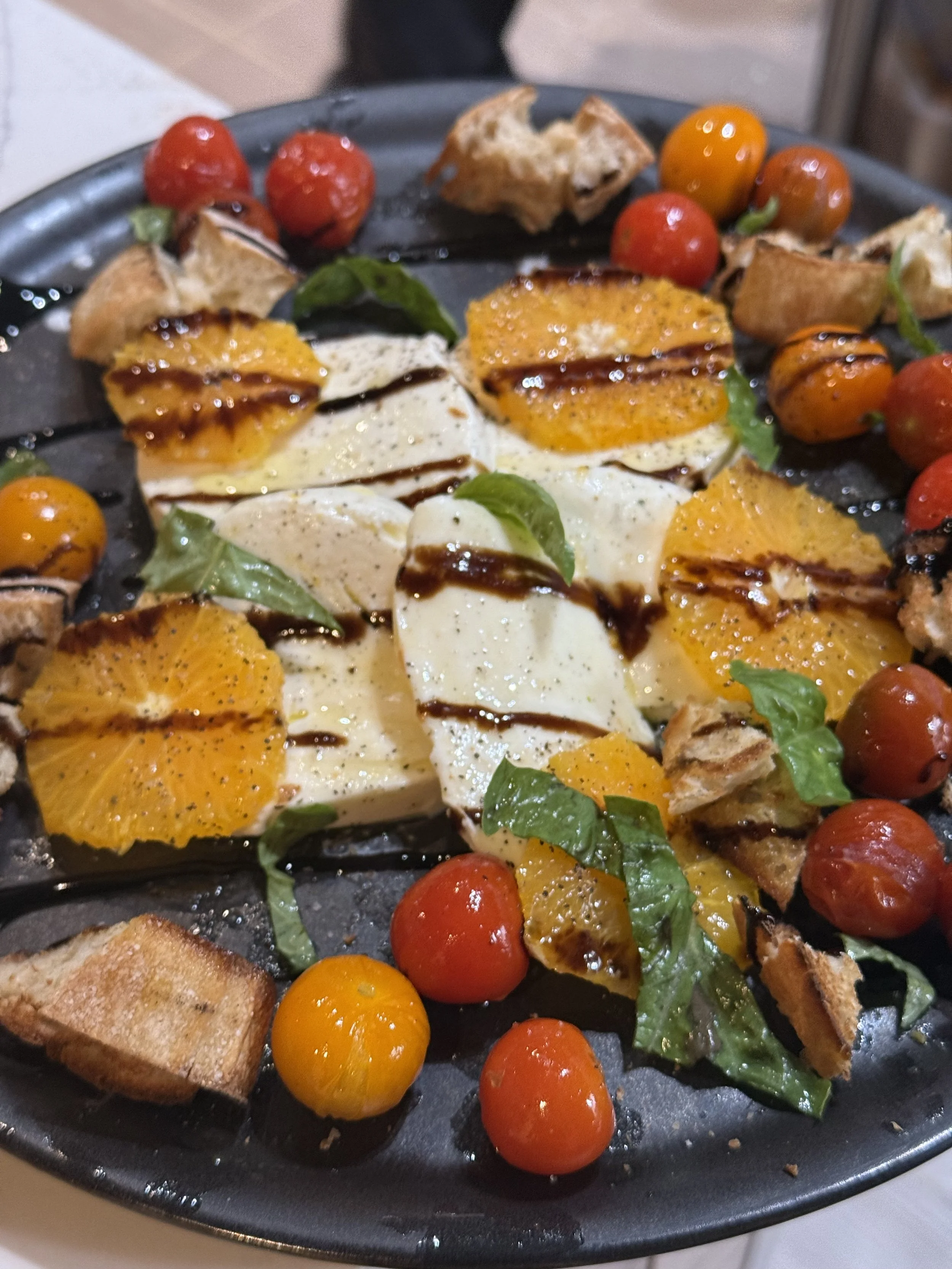 Fresh Caprese salad with slices of mozzarella cheese, orange and cherry tomatoes, basil leaves, balsamic glaze, toasted bread cubes, and greens on a black serving platter.
