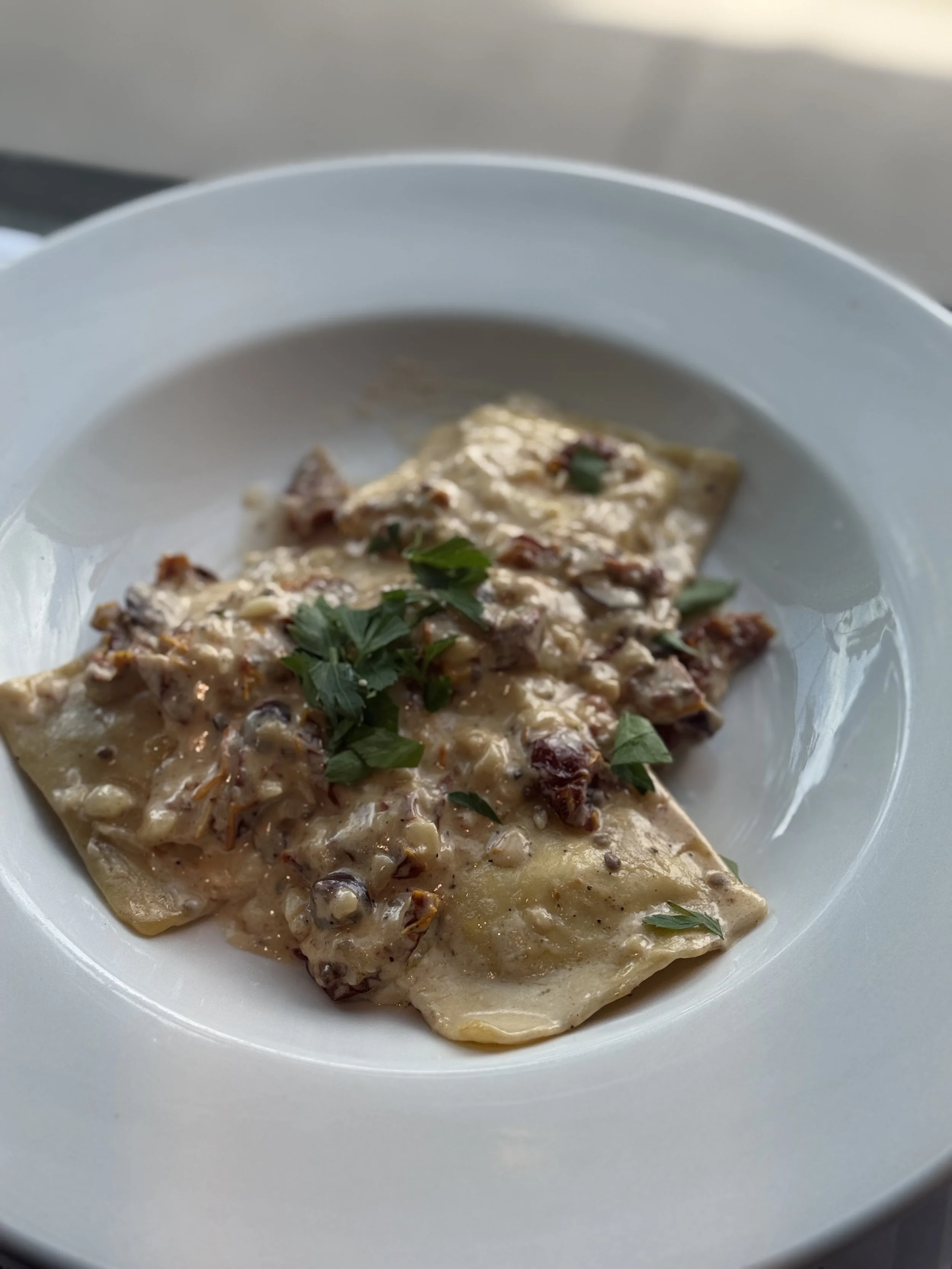 A white bowl with ravioli pasta topped with a creamy sauce, chopped herbs, and bits of meat.