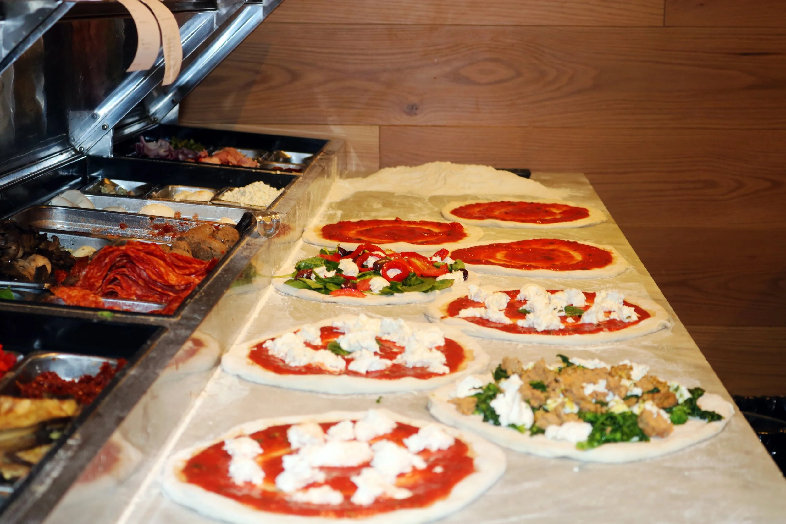  Pizza dough with tomato sauce, cheese, vegetables, and toppings on a wooden counter, with ingredients and toppings in a metal container to the left.