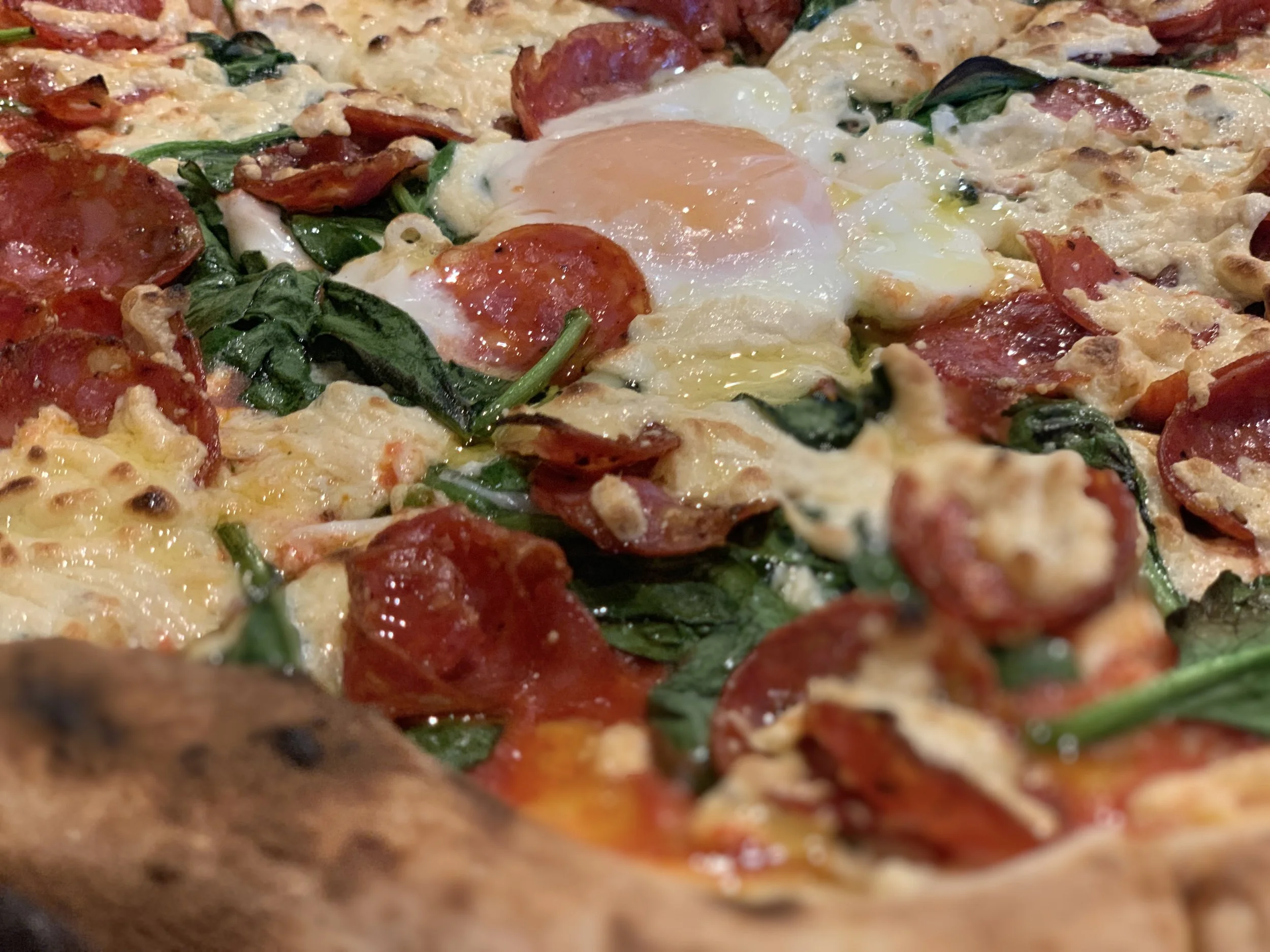 Close-up of a pizza with pepperoni, spinach, cheese, and a Soft cooked egg in the center.