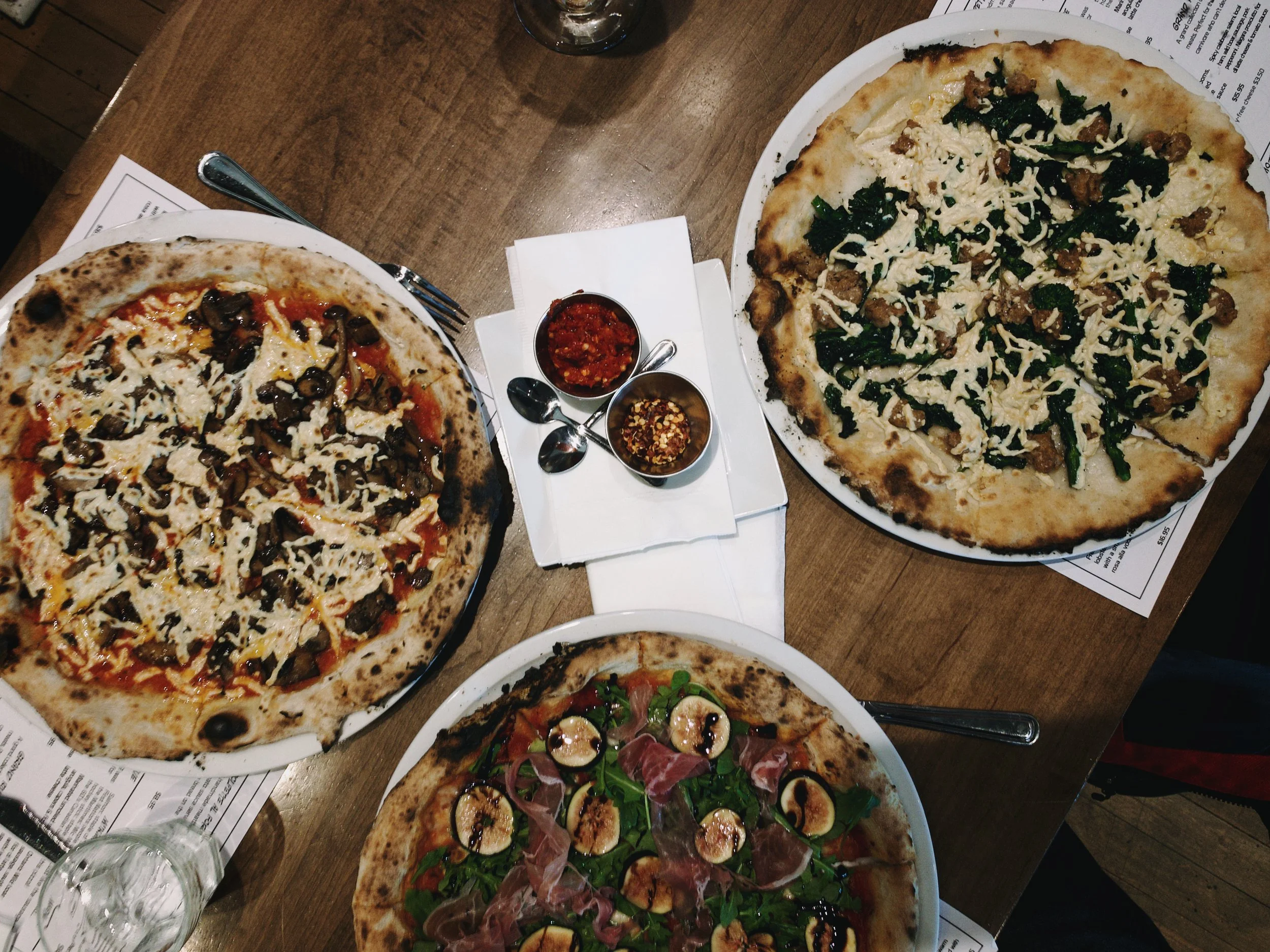 Three pizzas on a wooden table with two small bowls of dipping sauces in the center, one pizza topped with mushrooms, cheese, and tomato sauce, another with leafy greens, cheese, and sausage, and the third with prosciutto, figs, greens, and cheese.