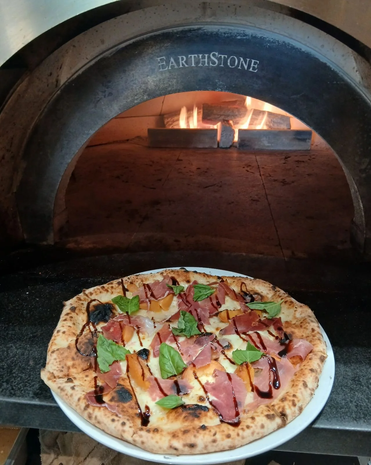 Pizza with ham, basil, peach slices, and balsamic glaze baking in a wood-fired oven.