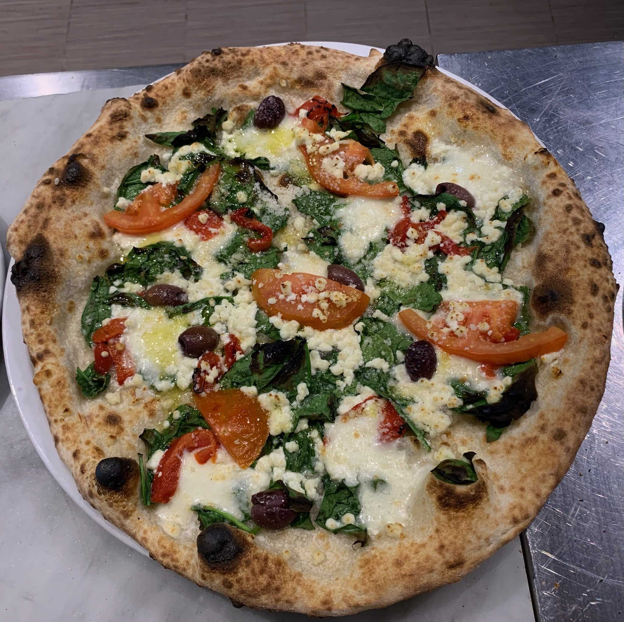 A pizza topped with cherry tomatoes, black olives, spinach, red bell peppers, and crumbled cheese on a golden-brown crust.