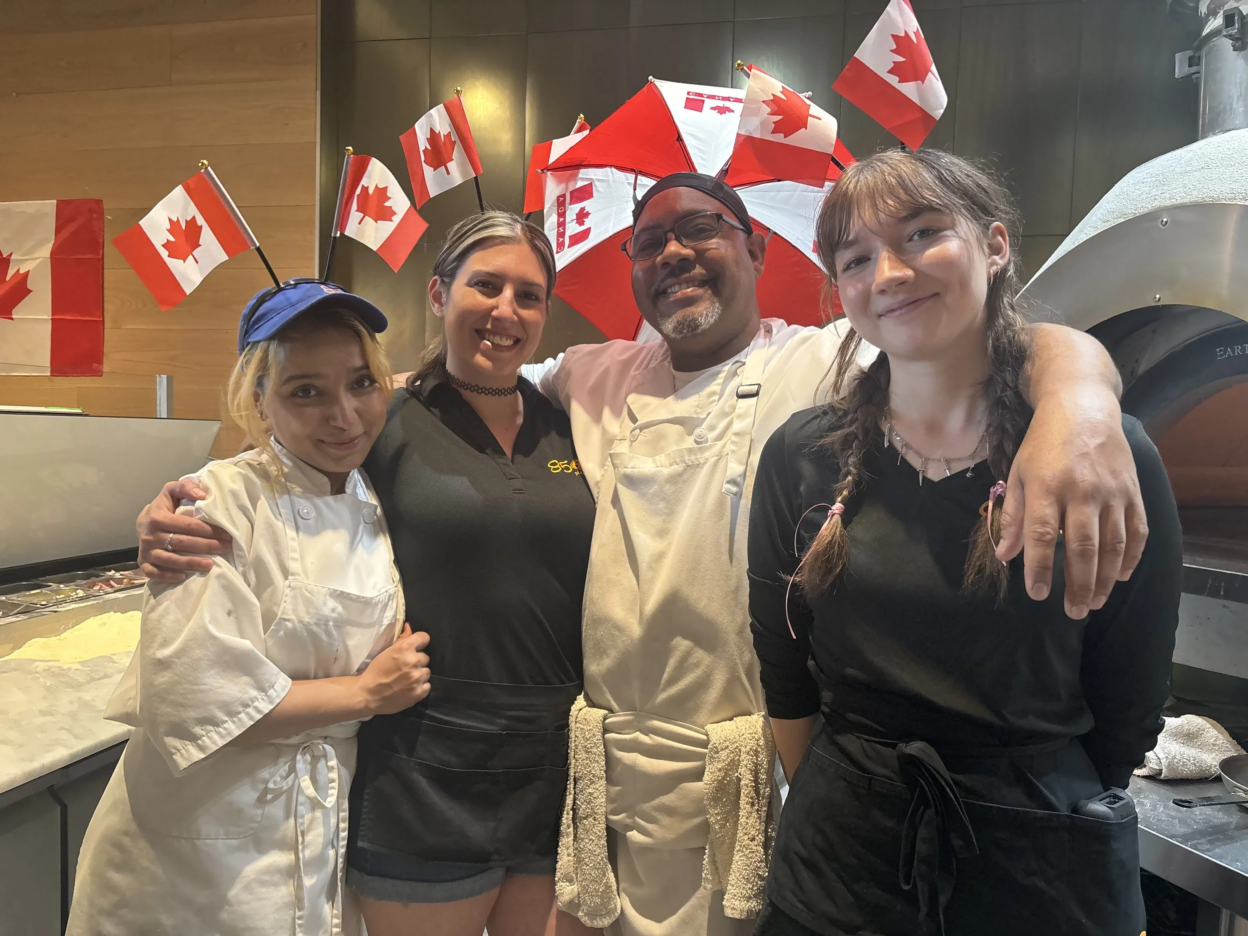 Four people wearing black aprons and a chef's coat pose together in a kitchen decorated with Canadian flags. One person has small Canadian flags on headbands, and a red and white umbrella with the Canadian flag pattern is behind them, all smiling at 