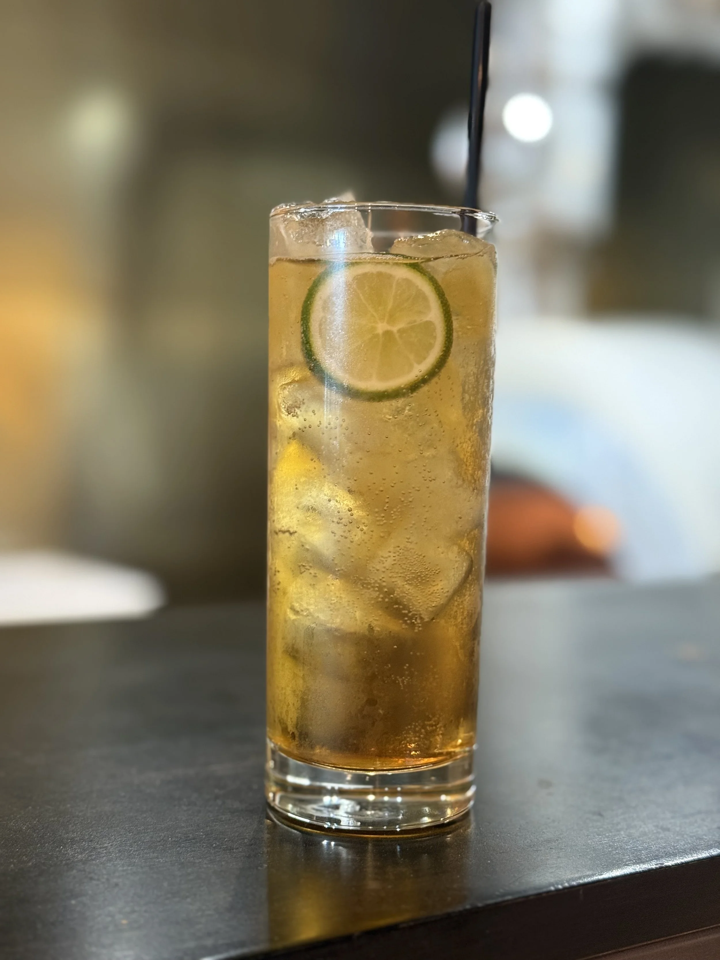 A tall glass of iced tea with ice cubes and a slice of lime, with a black straw in a blurred indoor setting.