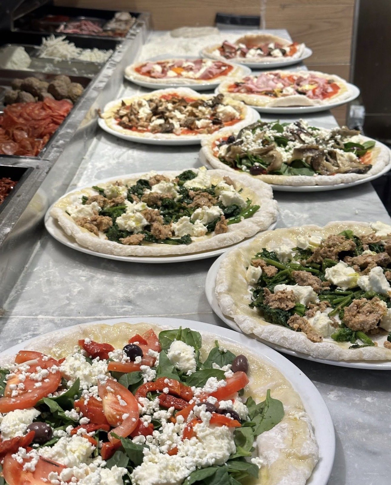 Several uncooked pizzas with various toppings including tomatoes, spinach, cheese, and meats, lined up on a table.