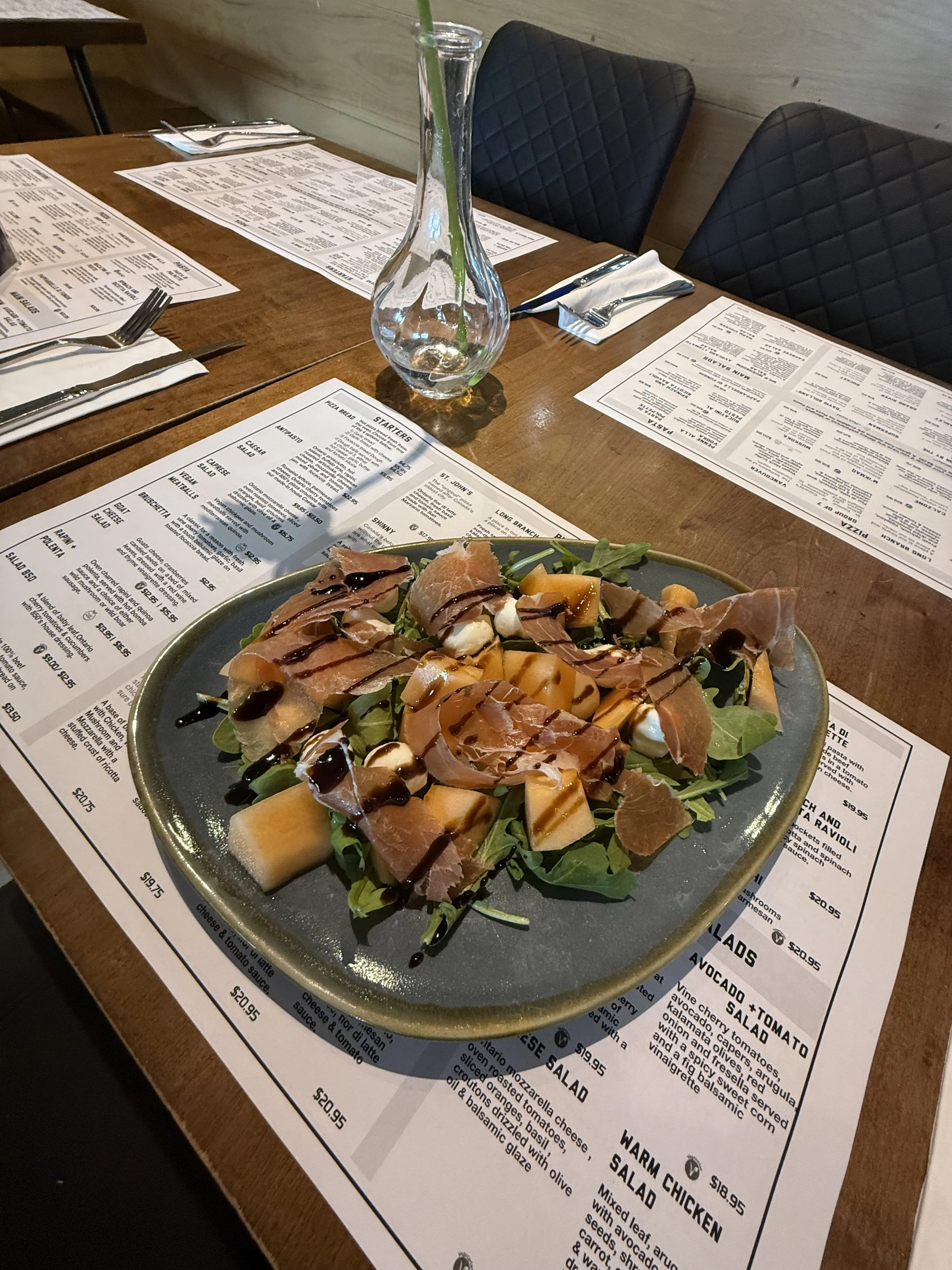 A salad with greens, cantaloupe, prosciutto, mozzarella cheese, and balsamic drizzle on a dark gray plate on a restaurant table.