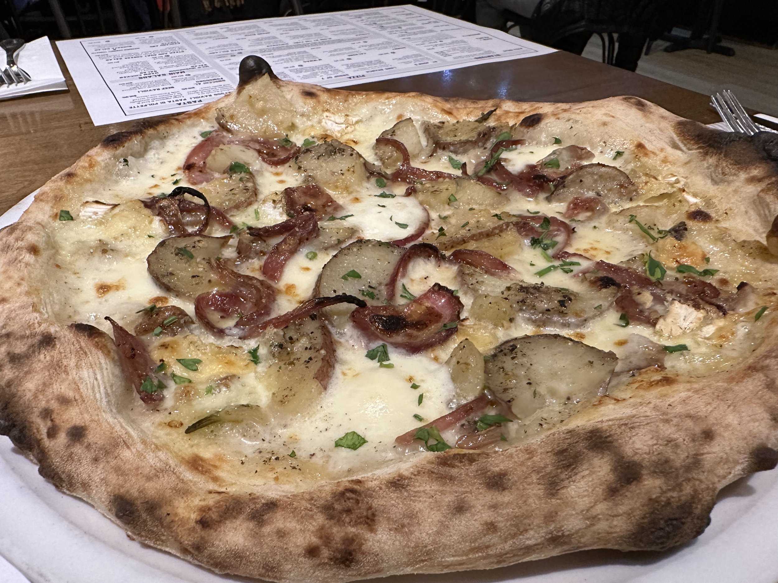 A rectangular pizza with cheese, potato slices, bacon, and green herbs on a wooden table.