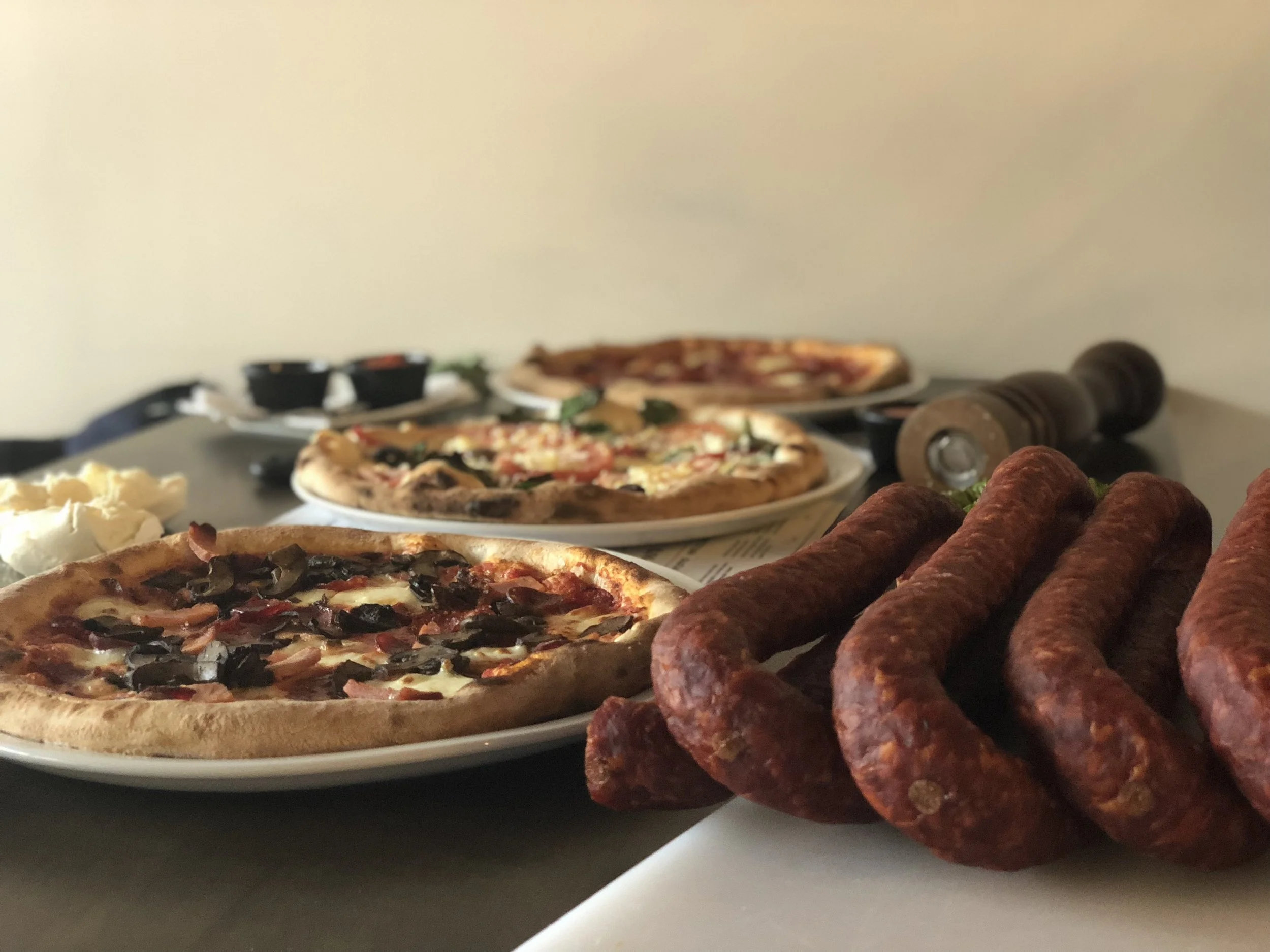 Plate of cooked sausages in the foreground and assorted pizzas on plates in the background, with dipping sauces and a pepper grinder on the table.