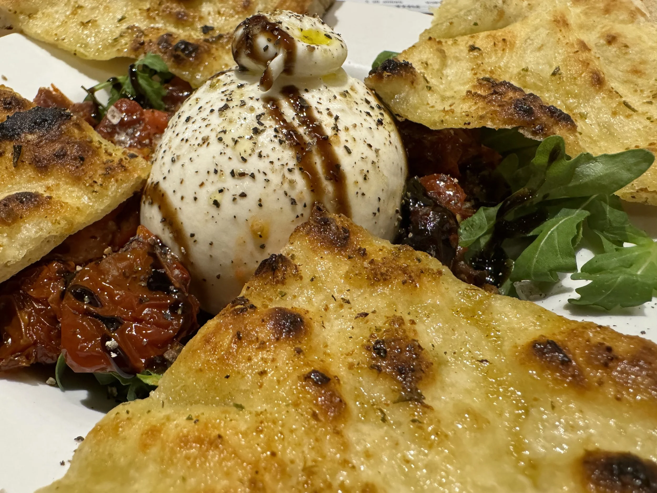 Close-up of a burrata cheese ball topped with balsamic glaze, surrounded by fried bread triangles, sun-dried tomatoes, and fresh arugula leaves.