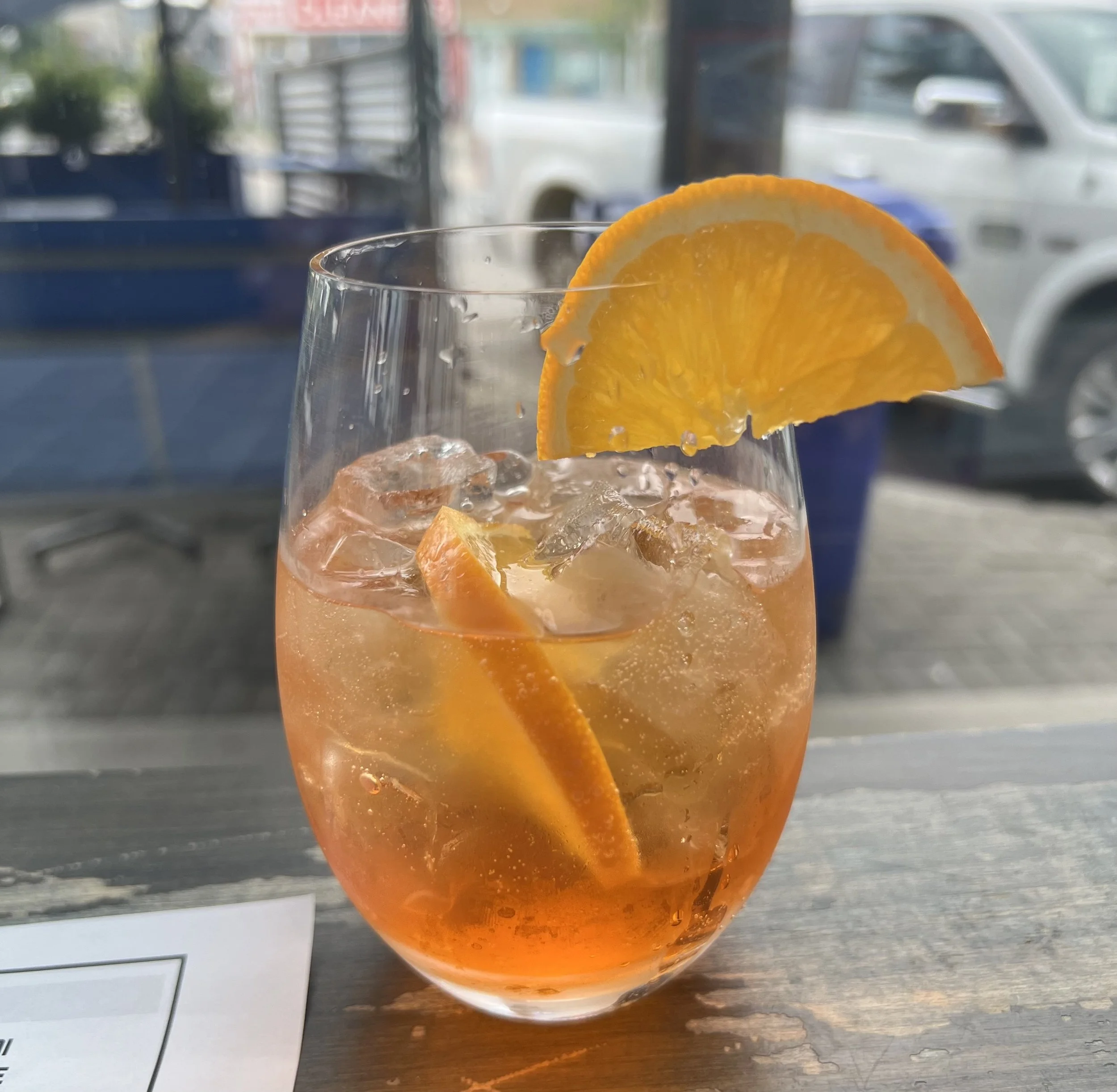 A glass of iced Aperol beverage garnished with a slice of orange, sitting on a wooden surface near a window with a street scene in the background.