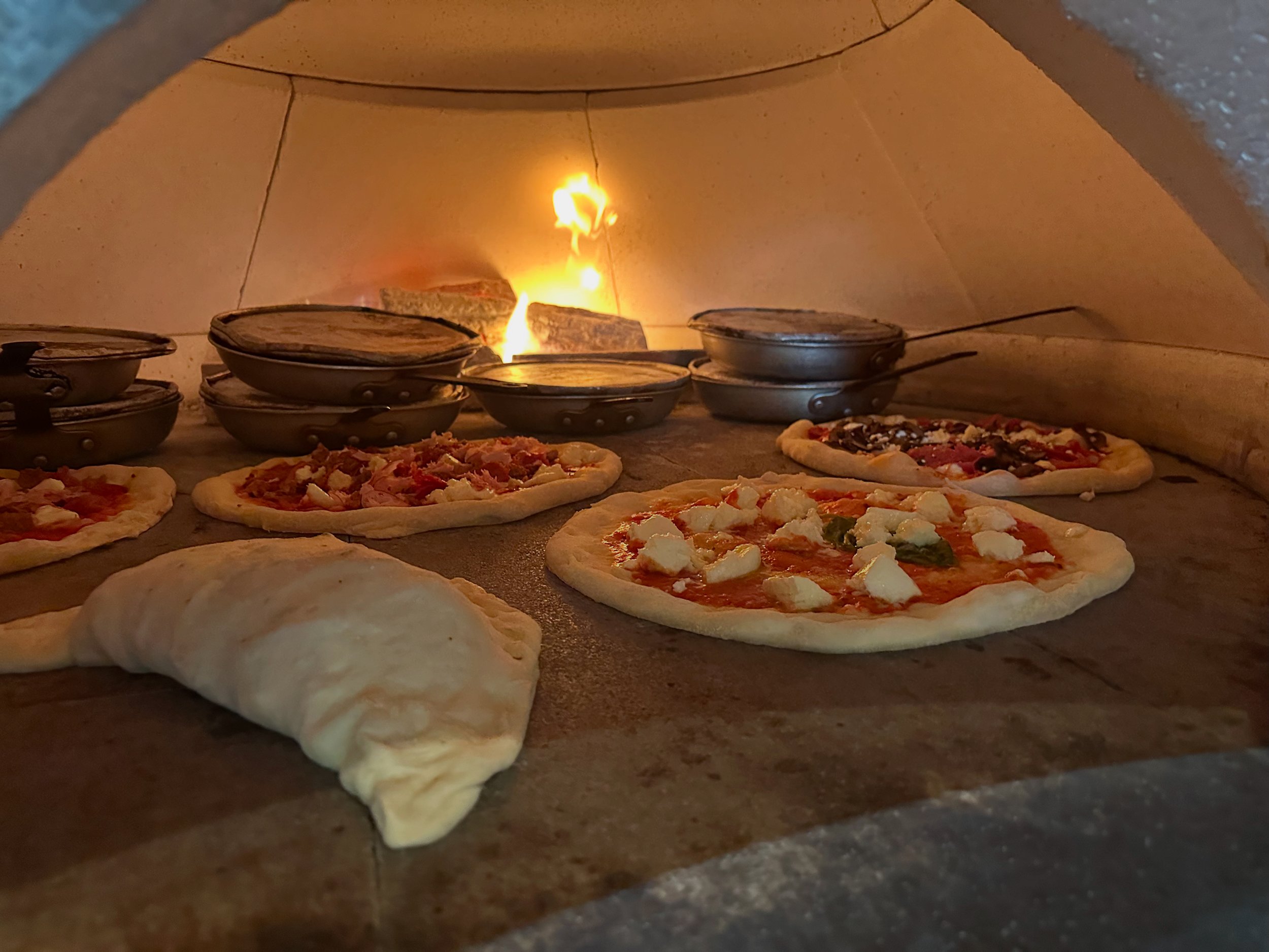 Pizzas baking inside a wood-fired oven with flames visible in the background.
