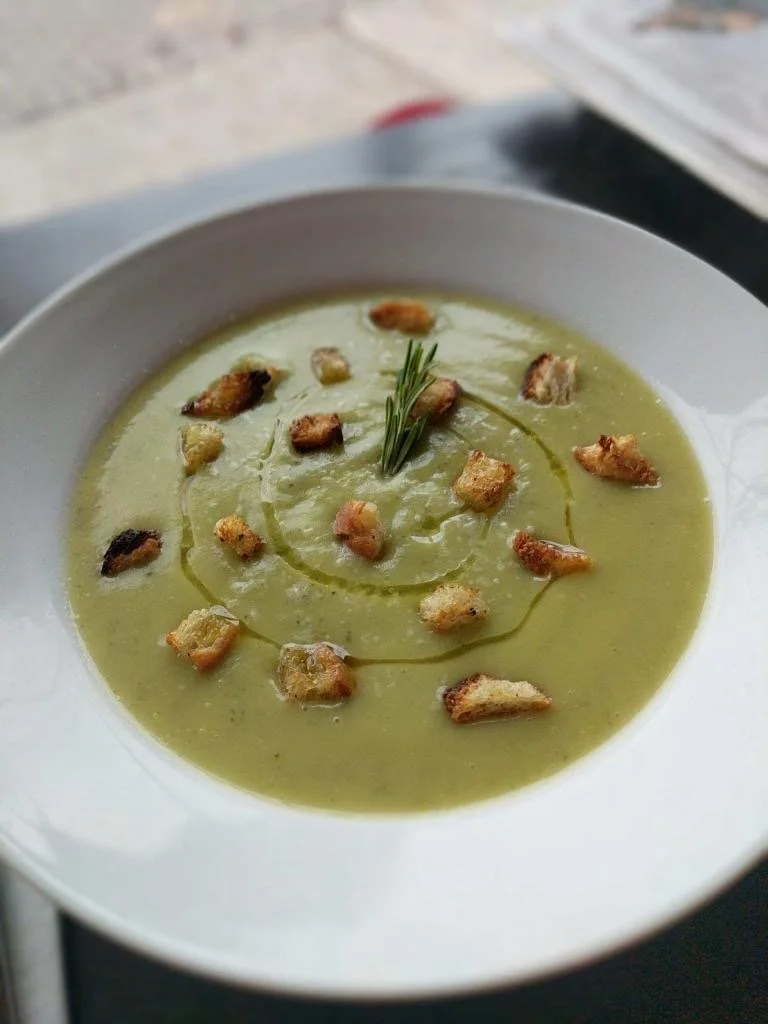 A bowl of green soup garnished with croutons and a sprig of rosemary.