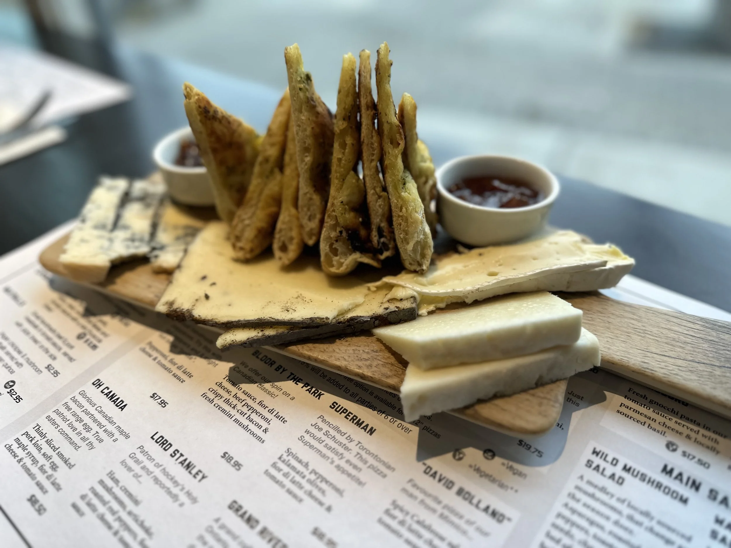 A wooden platter with assorted cheeses, crispy breadsticks, and two small bowls of dipping sauce, placed on a napkin-covered table.