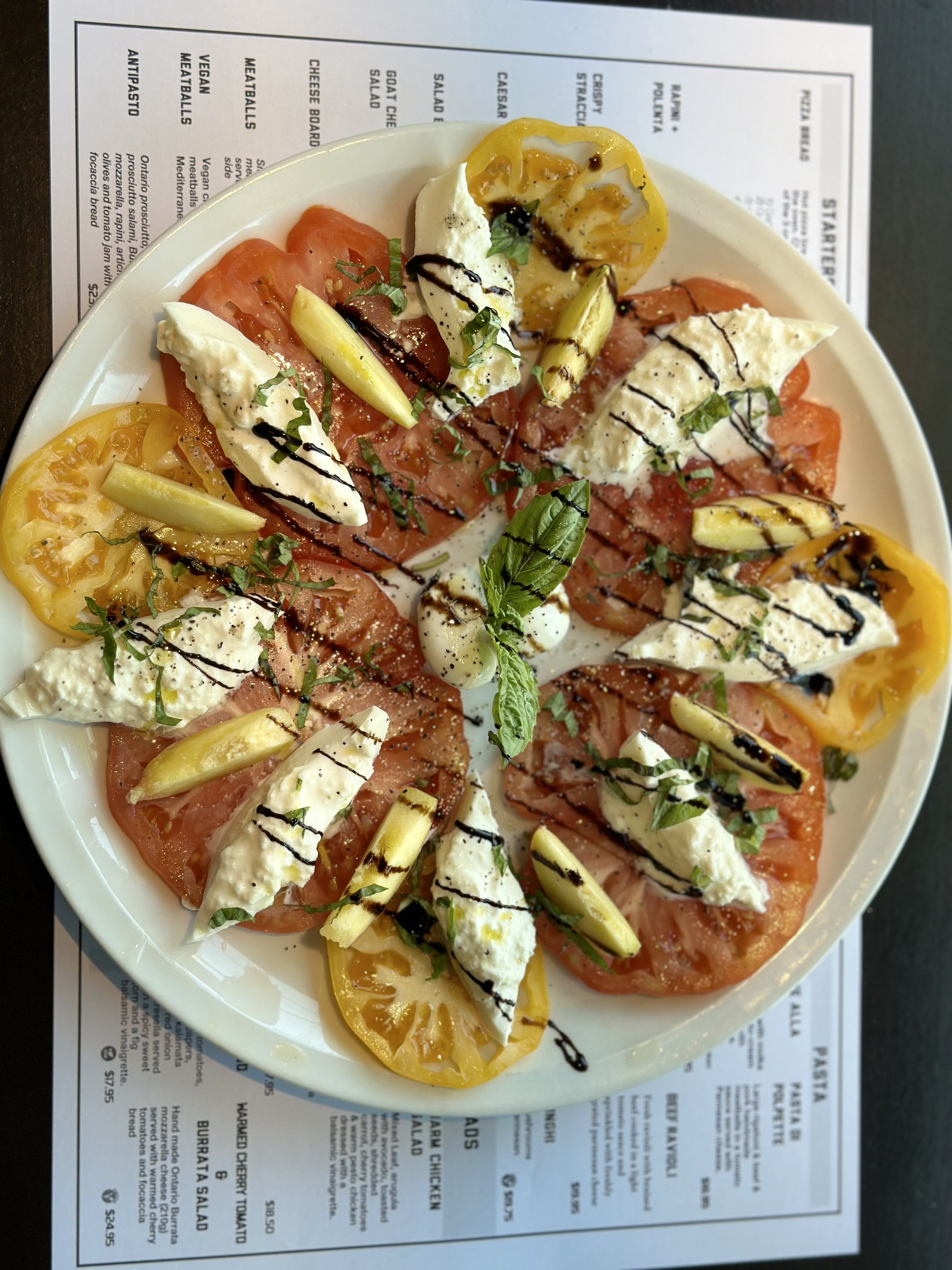 Plate of sliced tomatoes, fresh mozzarella cheese, yellow and green vegetables, drizzled with balsamic glaze, garnished with chopped herbs and a basil leaf, on a white plate with a menu underneath.