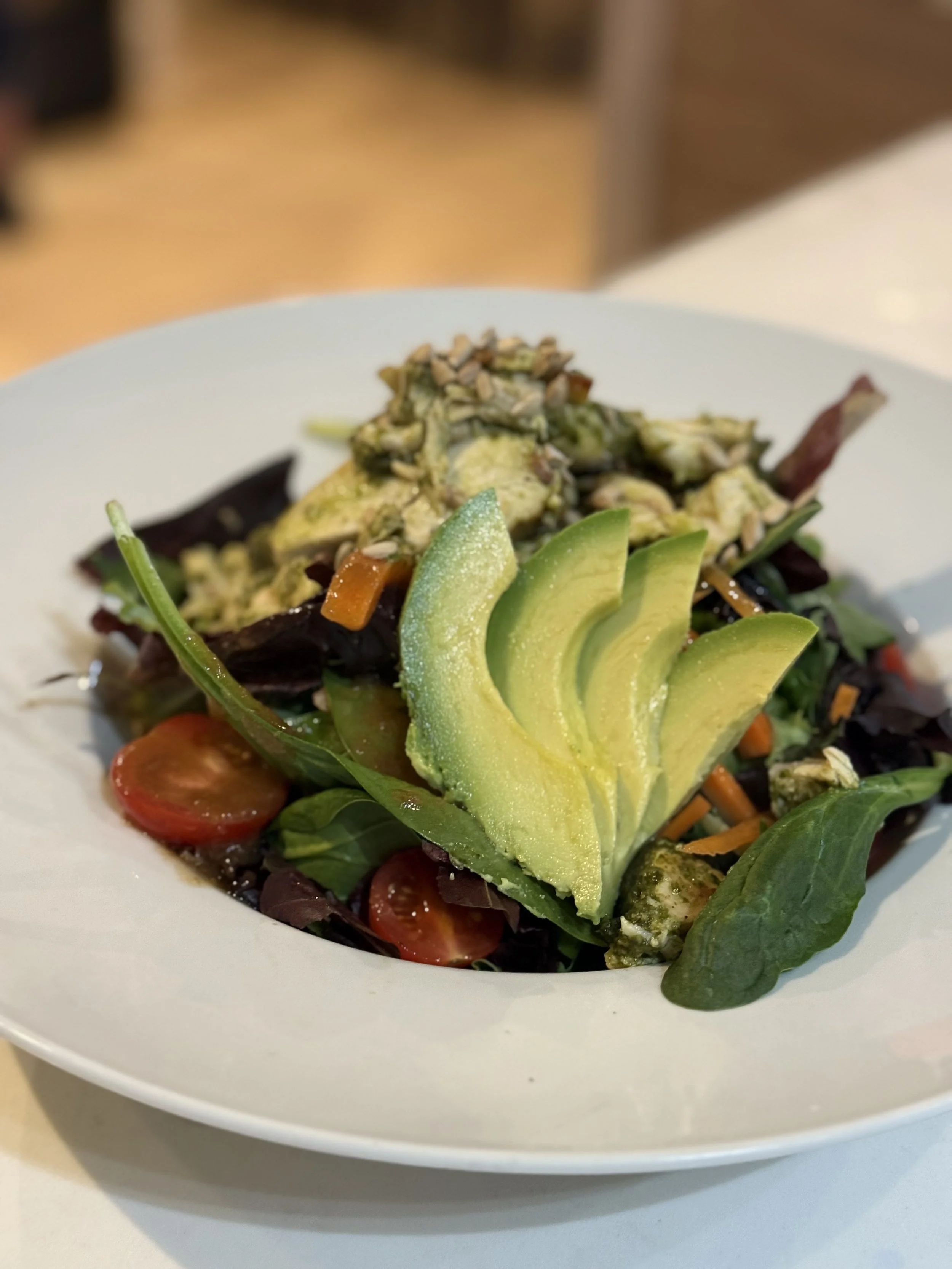 Fresh mixed greens salad with cherry tomatoes, sliced avocado, shredded carrots, sunflower seeds, and crumbled cheese on a white plate.