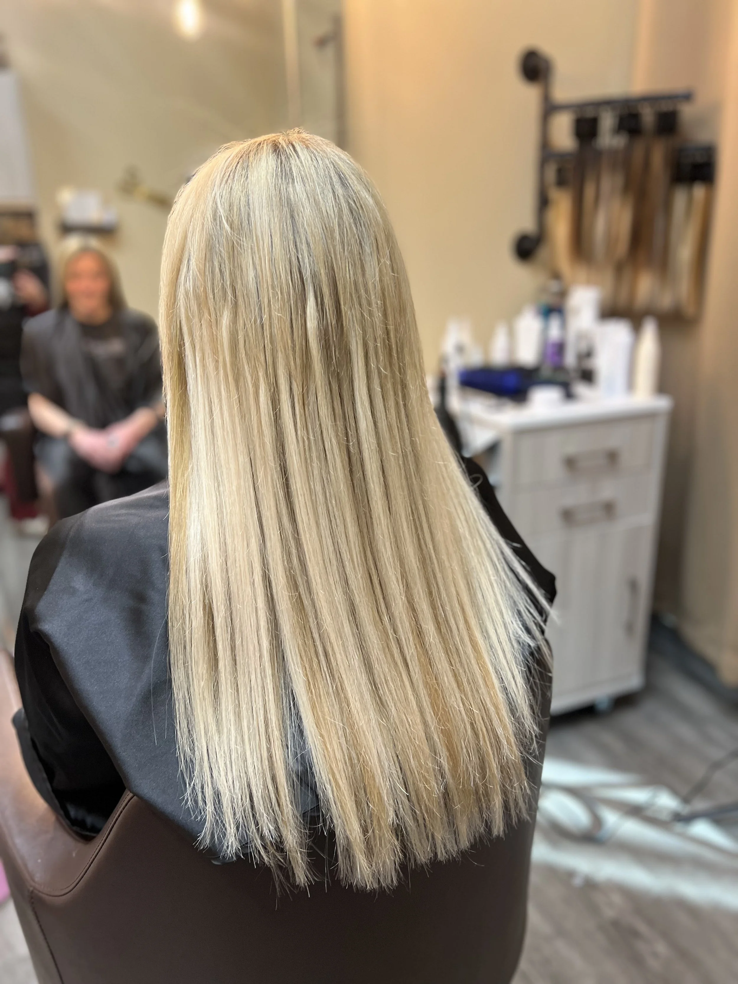 Blonde hair after hair coloring service at salon, with a person sitting in the background in a salon environment.