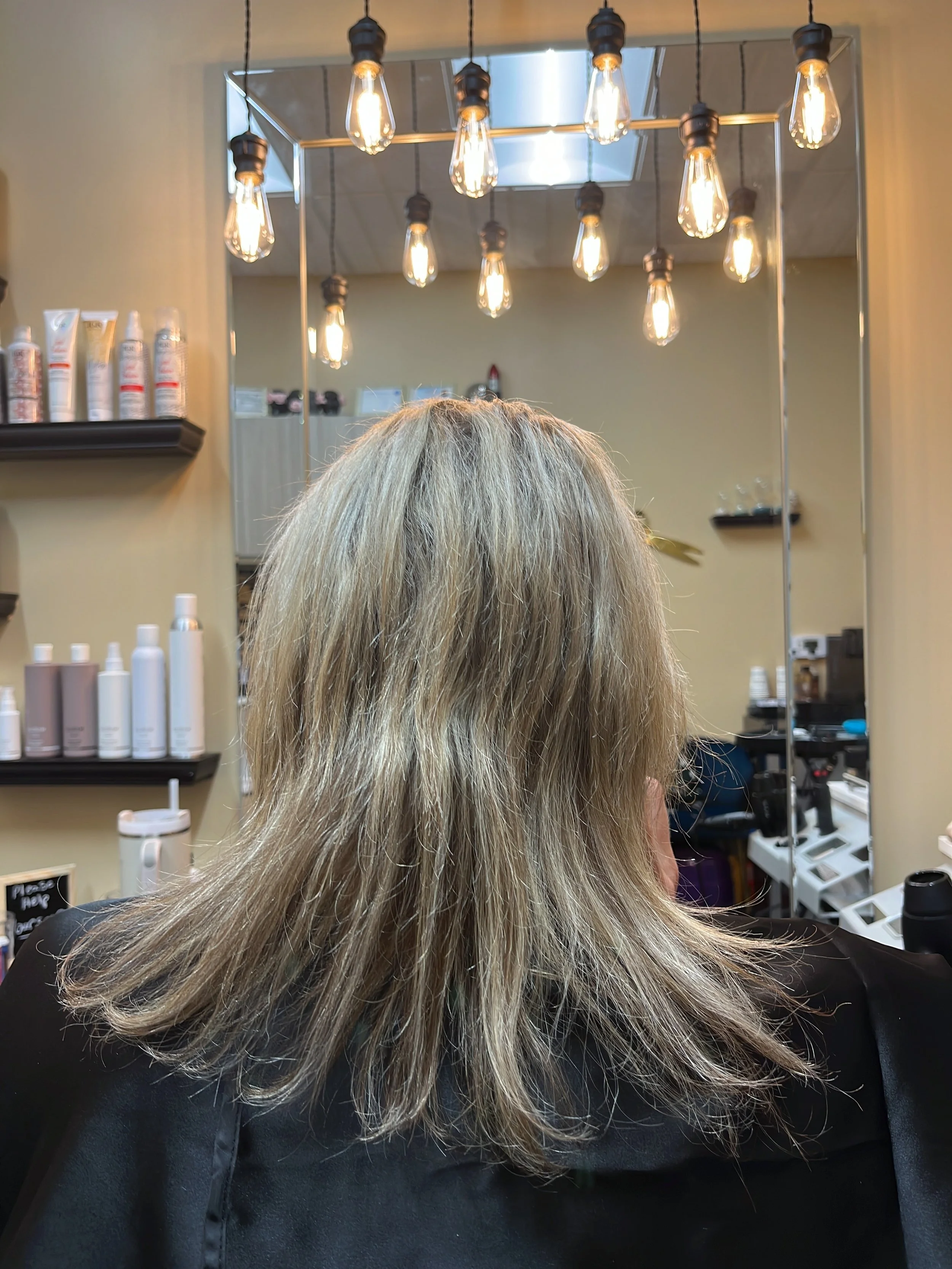 Back of a person's head with long, blonde hair sitting in a salon chair, facing a mirror with hanging Edison-style light bulbs and salon products visible in the background.