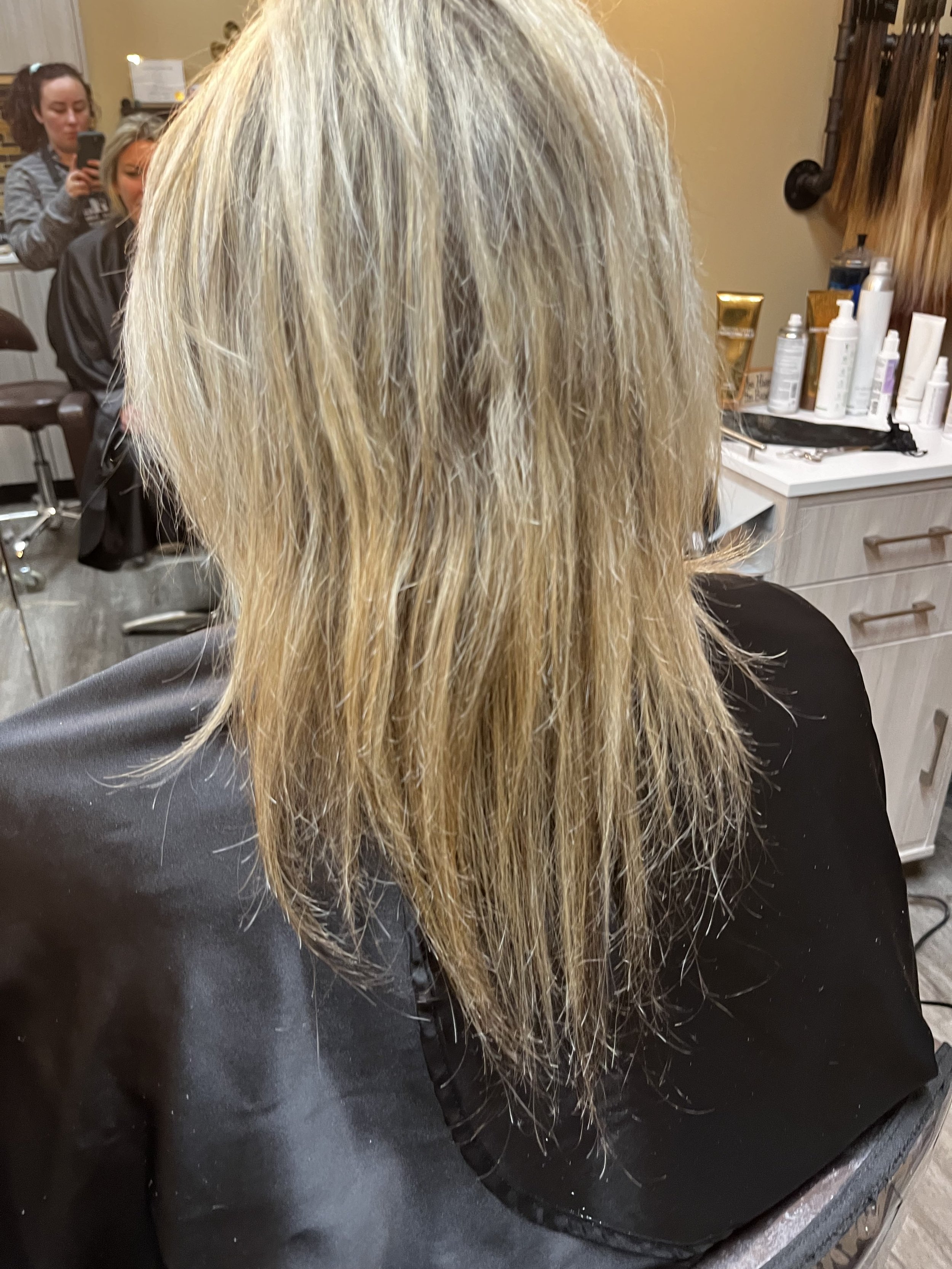 A person with long, blonde, wet hair sitting in a salon chair, facing away from the camera, with two women in the background, one taking a photo with a smartphone and the other watching. Salon bottles and tools are visible on a counter to the right.