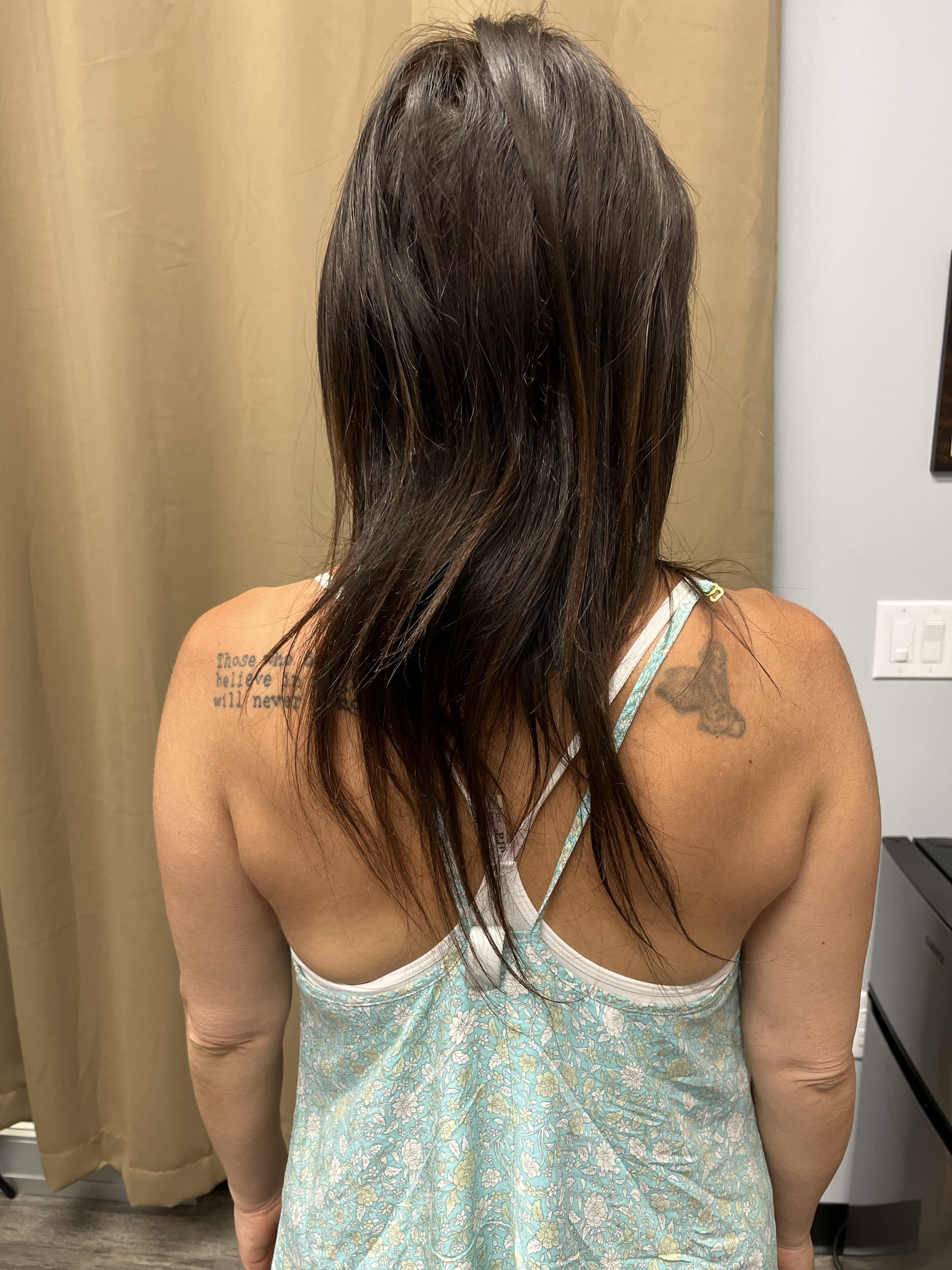 A woman with long brown hair standing indoors, wearing a light blue and white floral dress with thin straps. She has tattoos on her left shoulder and upper arm. The background shows a beige curtain and a portion of a white wall with a light switch.