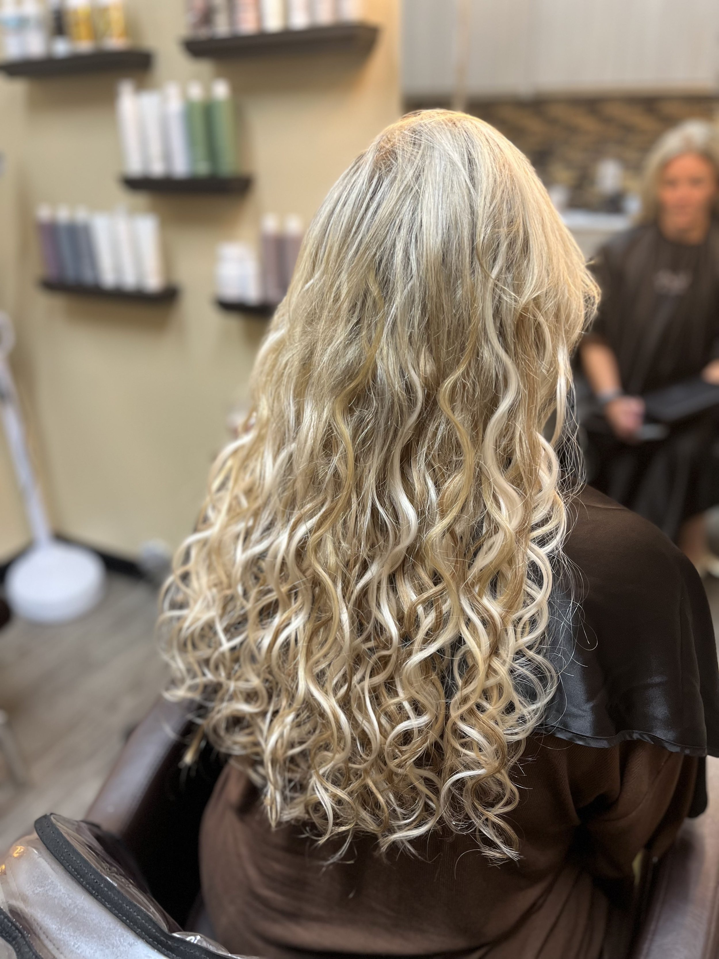 Back of a woman with long, blonde, curly hair sitting in salon chair, with a hairstylist in the background.