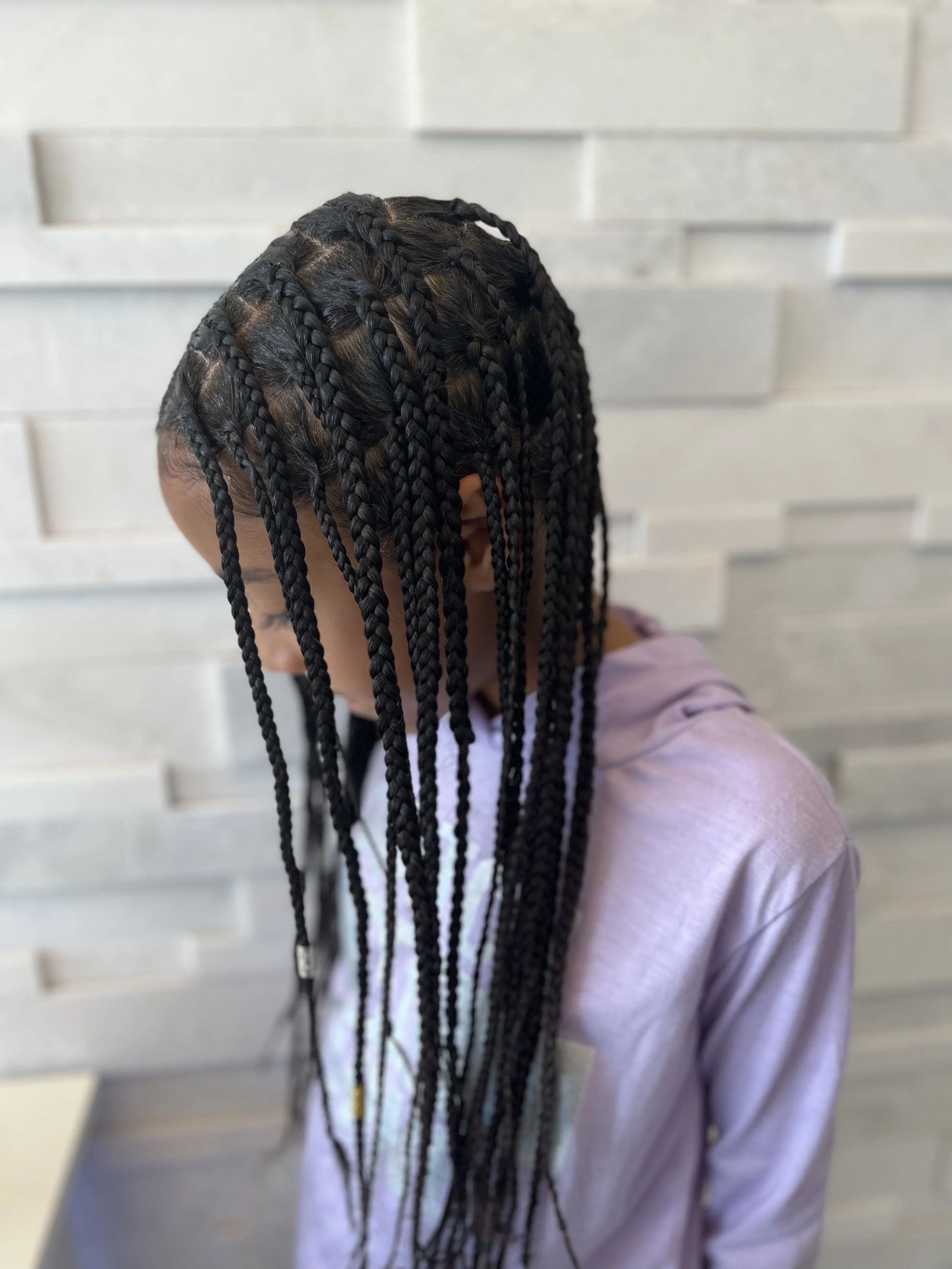Person with long box braids, wearing a light purple hoodie, standing against a textured gray wall.