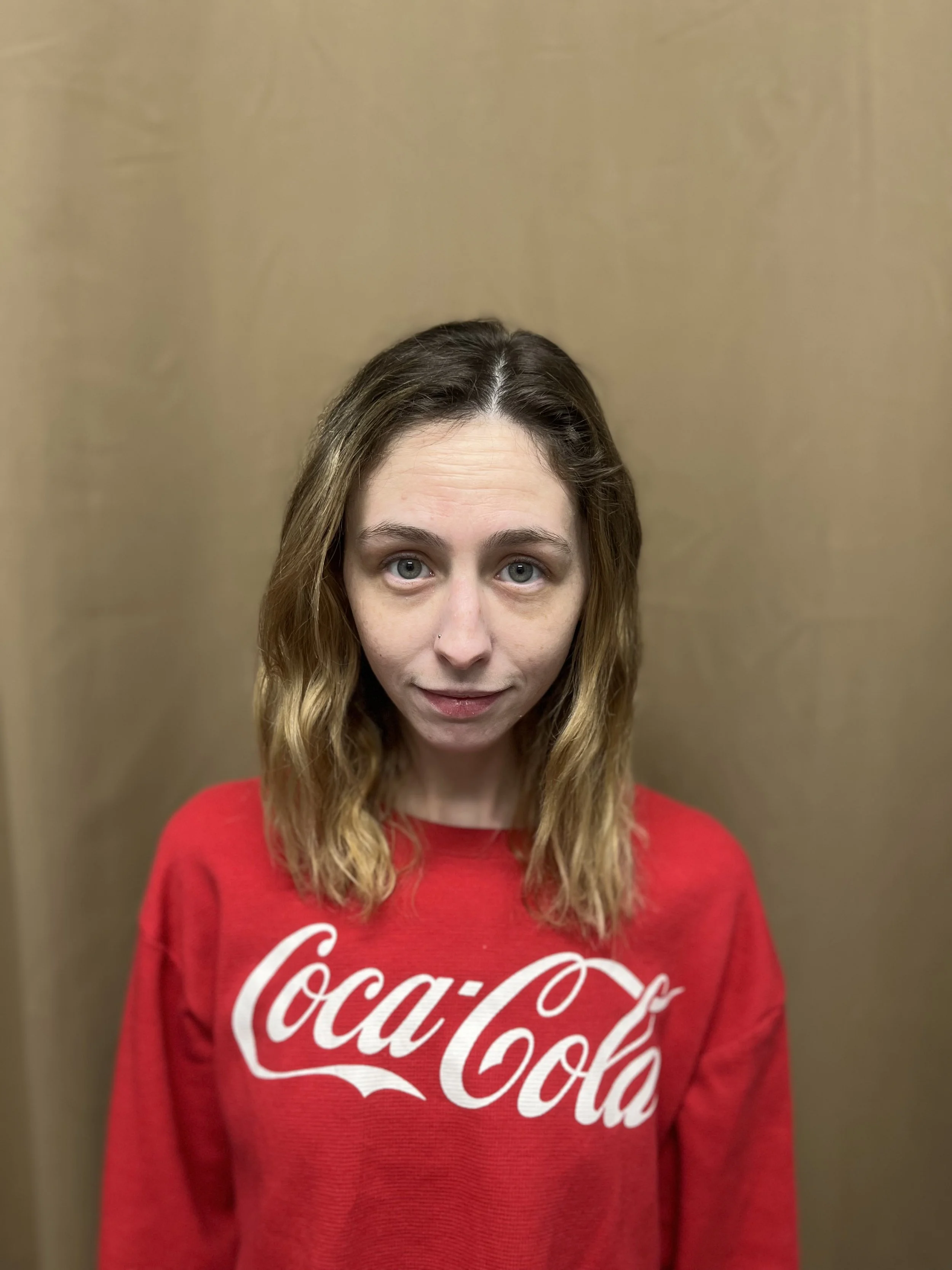 A woman with shoulder-length wavy hair and blue eyes wearing a red Coca-Cola sweatshirt standing against a plain beige background.