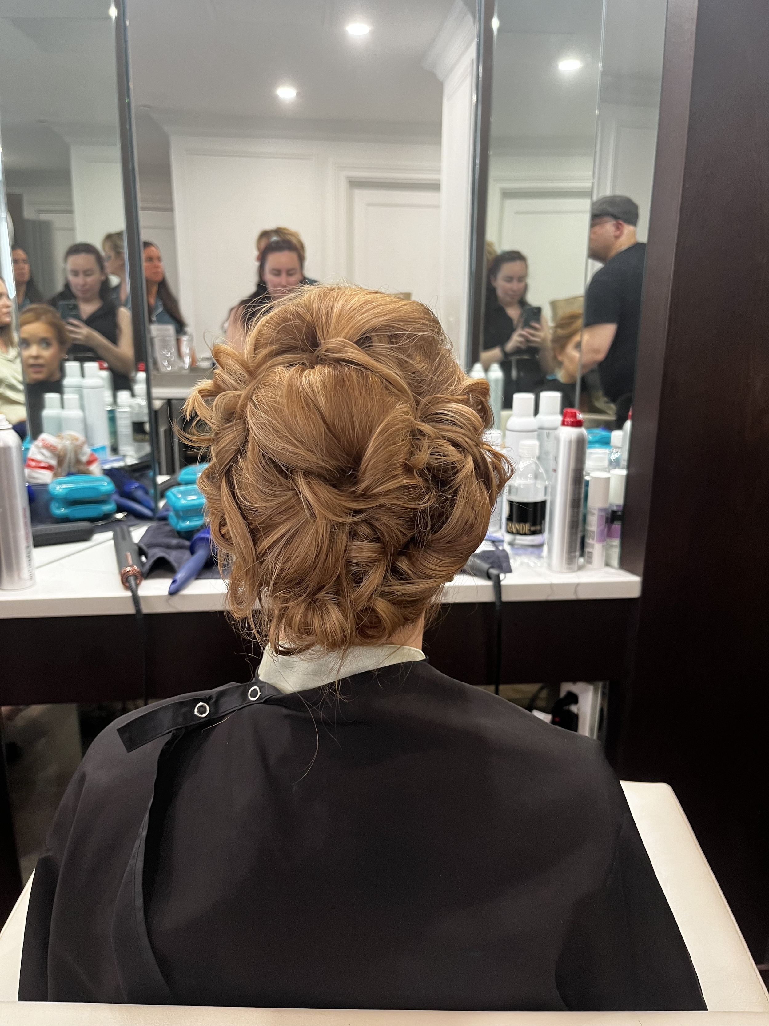 Woman with brown hair styled in an intricate updo sitting in a salon chair, with hairstyling products and people in the background.
