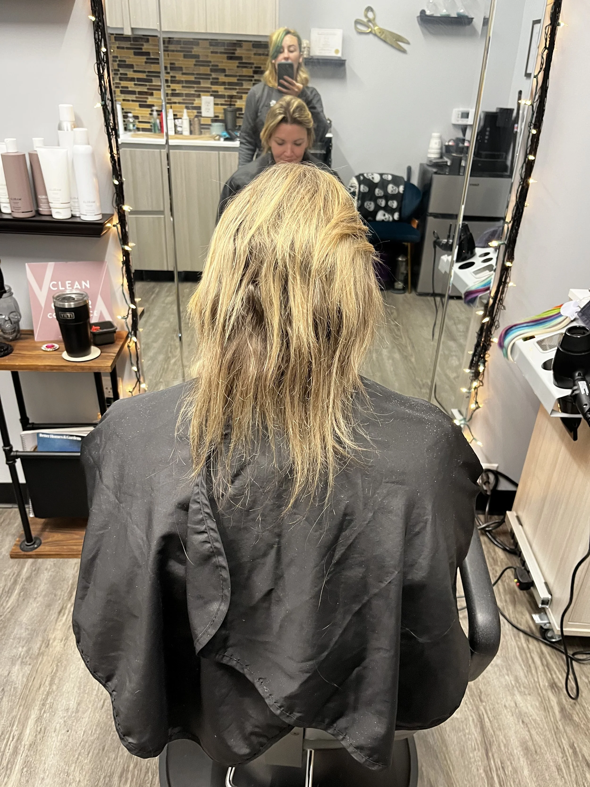 A woman with blonde hair sits in a salon chair with a black cape, facing a mirror. Two hairstylists are visible in the reflection, one taking a photo with a phone. Salon shelves with hair products and equipment are visible, along with decorations and