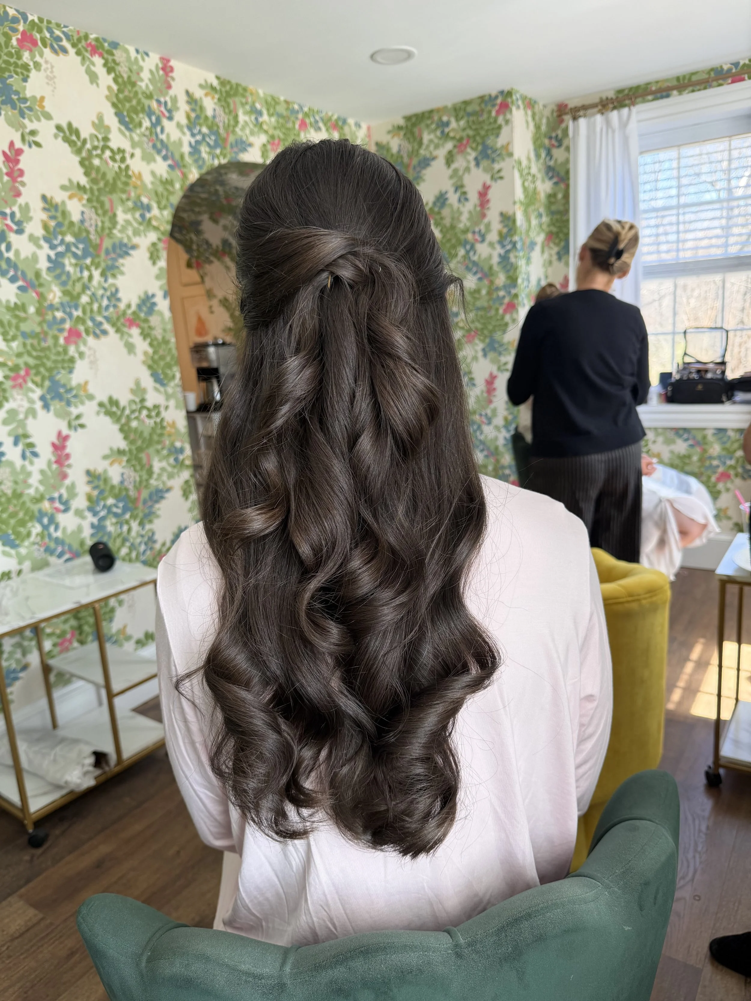 A woman with long, dark, curled hair styled in loose waves with a braid twist on top, sitting in a green armchair in a room with floral wallpaper, looking towards the window, with another woman in black standing near the window.