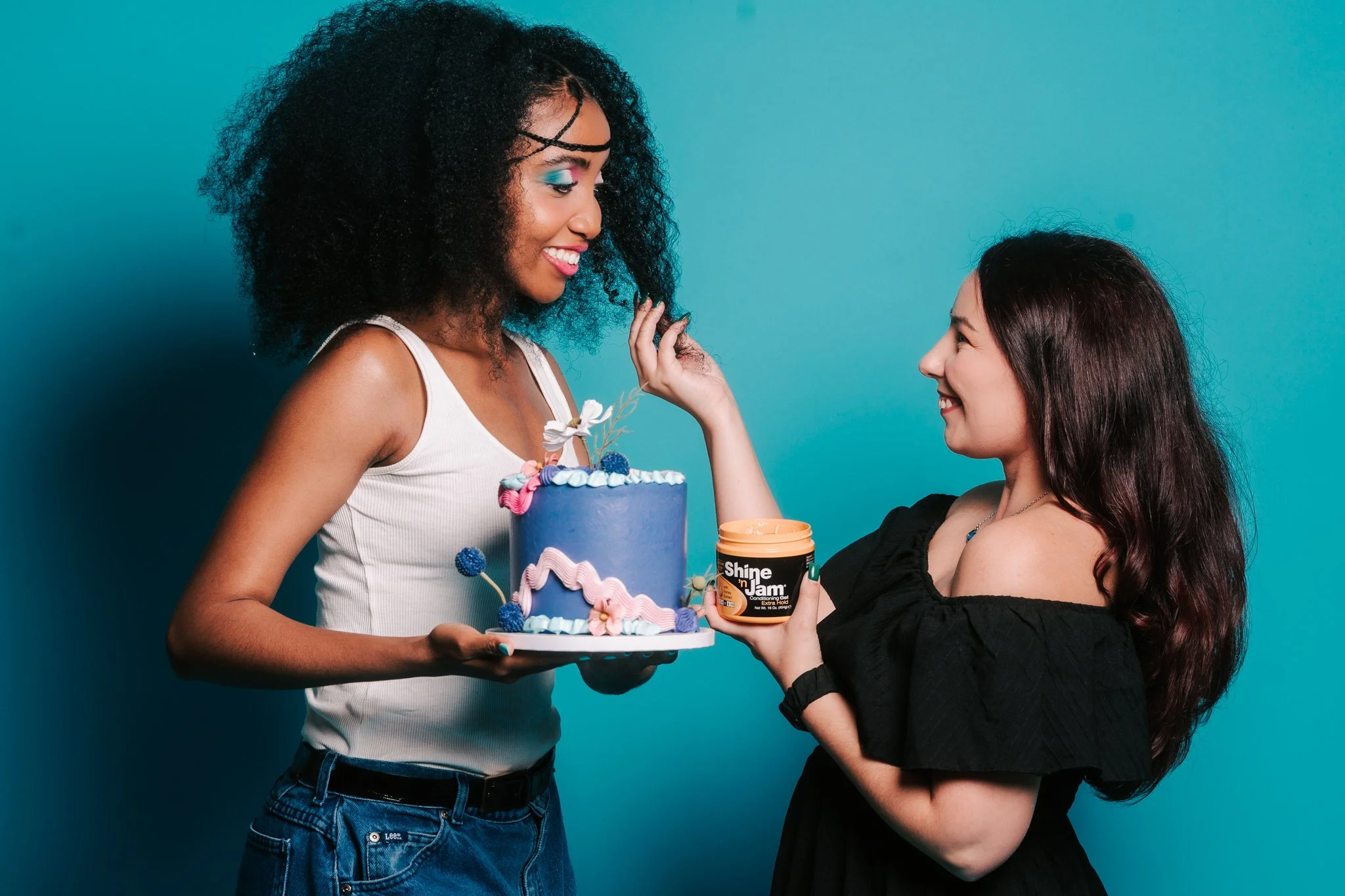 Two women smiling at each other against a turquoise background. One is holding a purple decorated cake, the other is holding a container labeled "Shine n Jam."