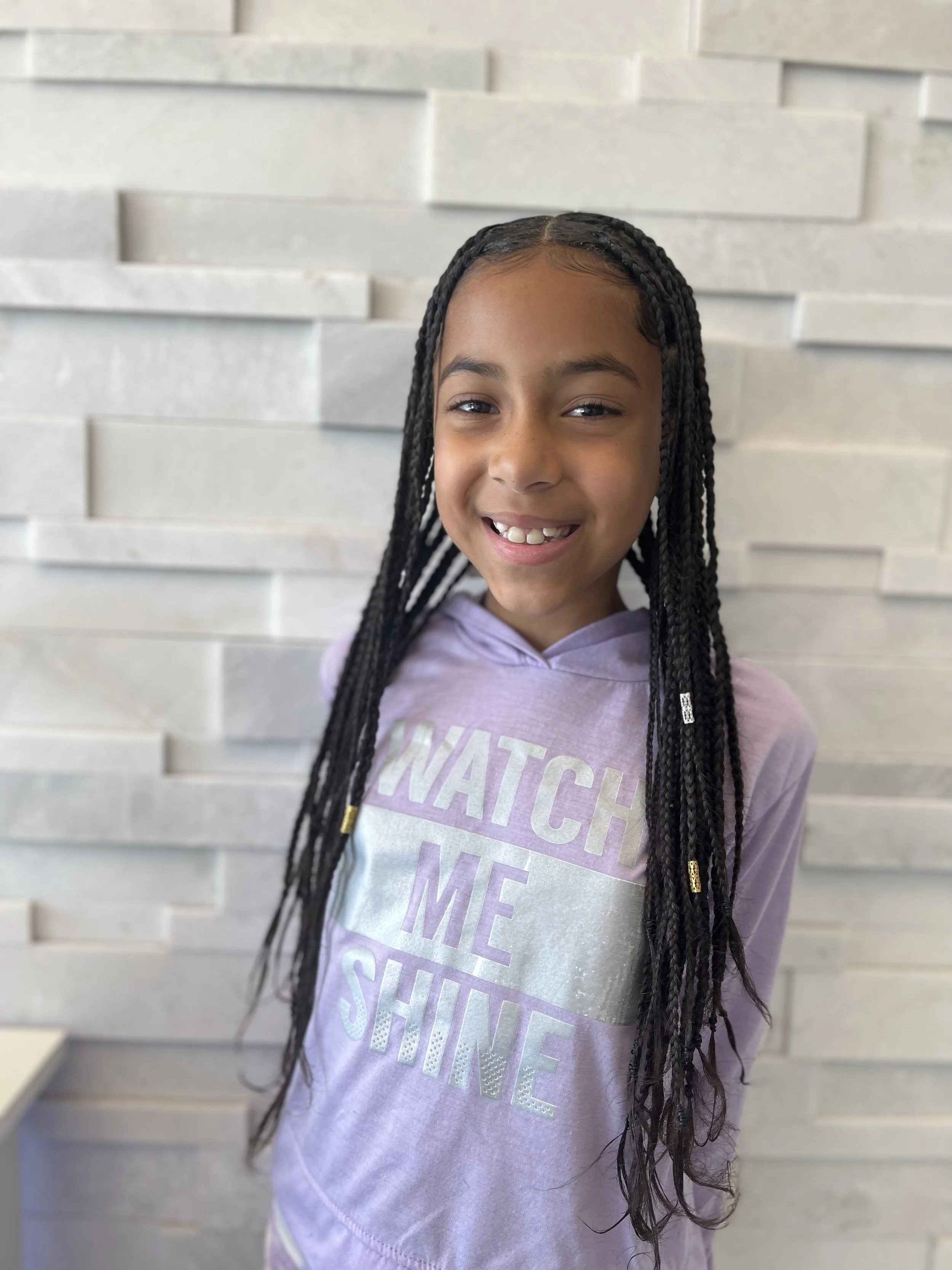 Smiling child with braided hair in box braids wearing a purple hoodie that says 'Watch Me Shine' against a textured stone wall background.