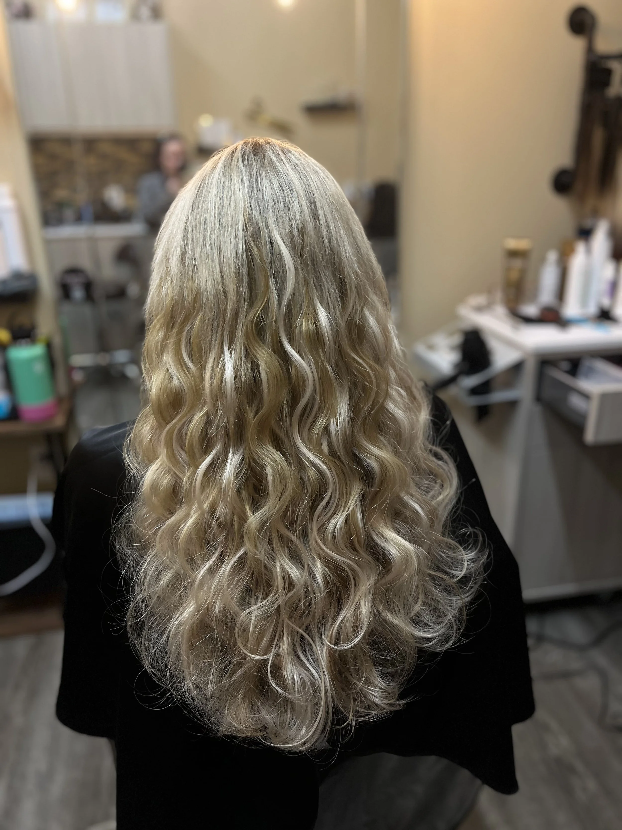 Back view of a person with long, curly, blonde hair in a salon or similar setting.