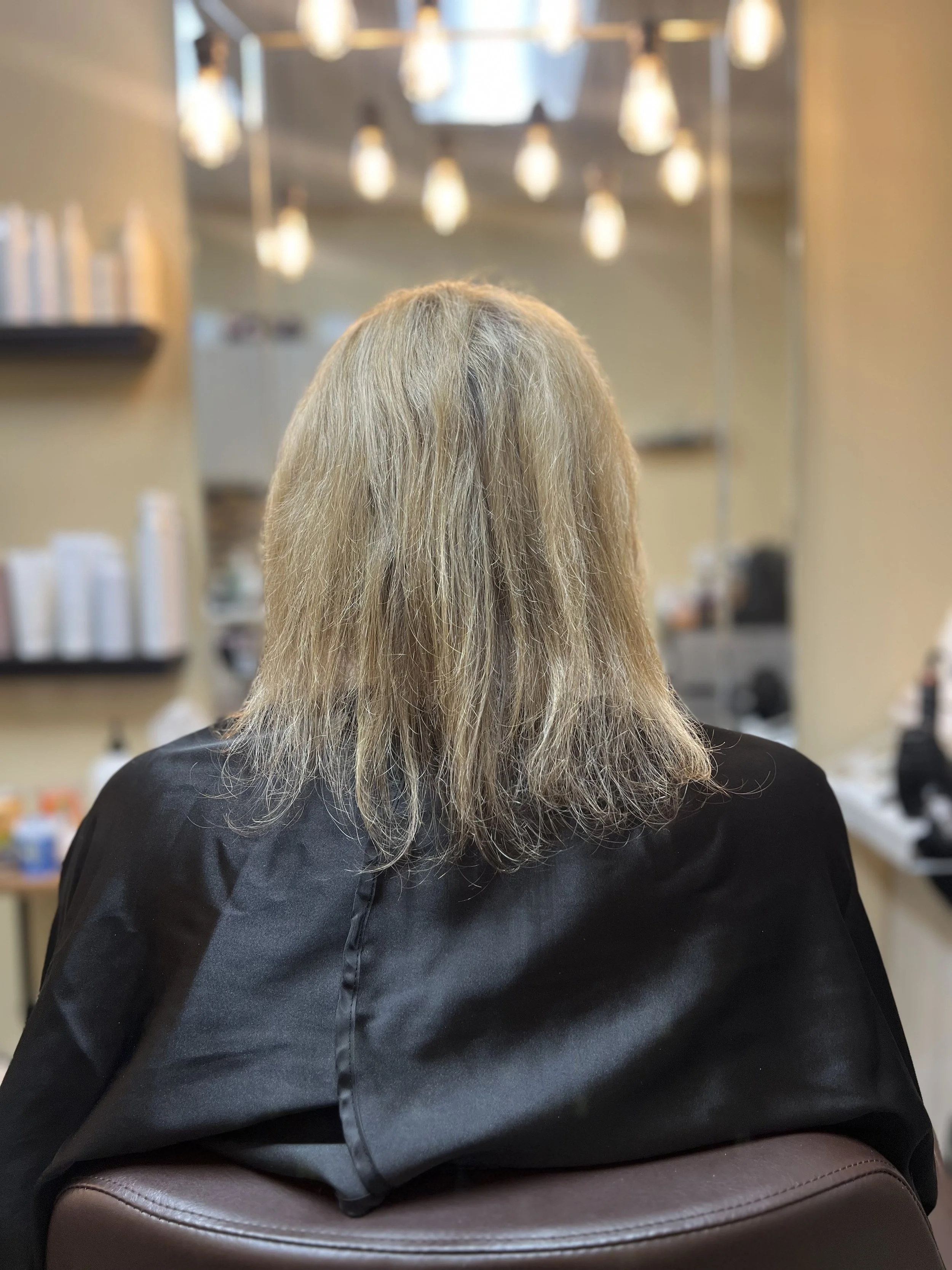 Back of a person with long, blond hair sitting in a salon chair, facing away, wearing a black cape at a hair salon.