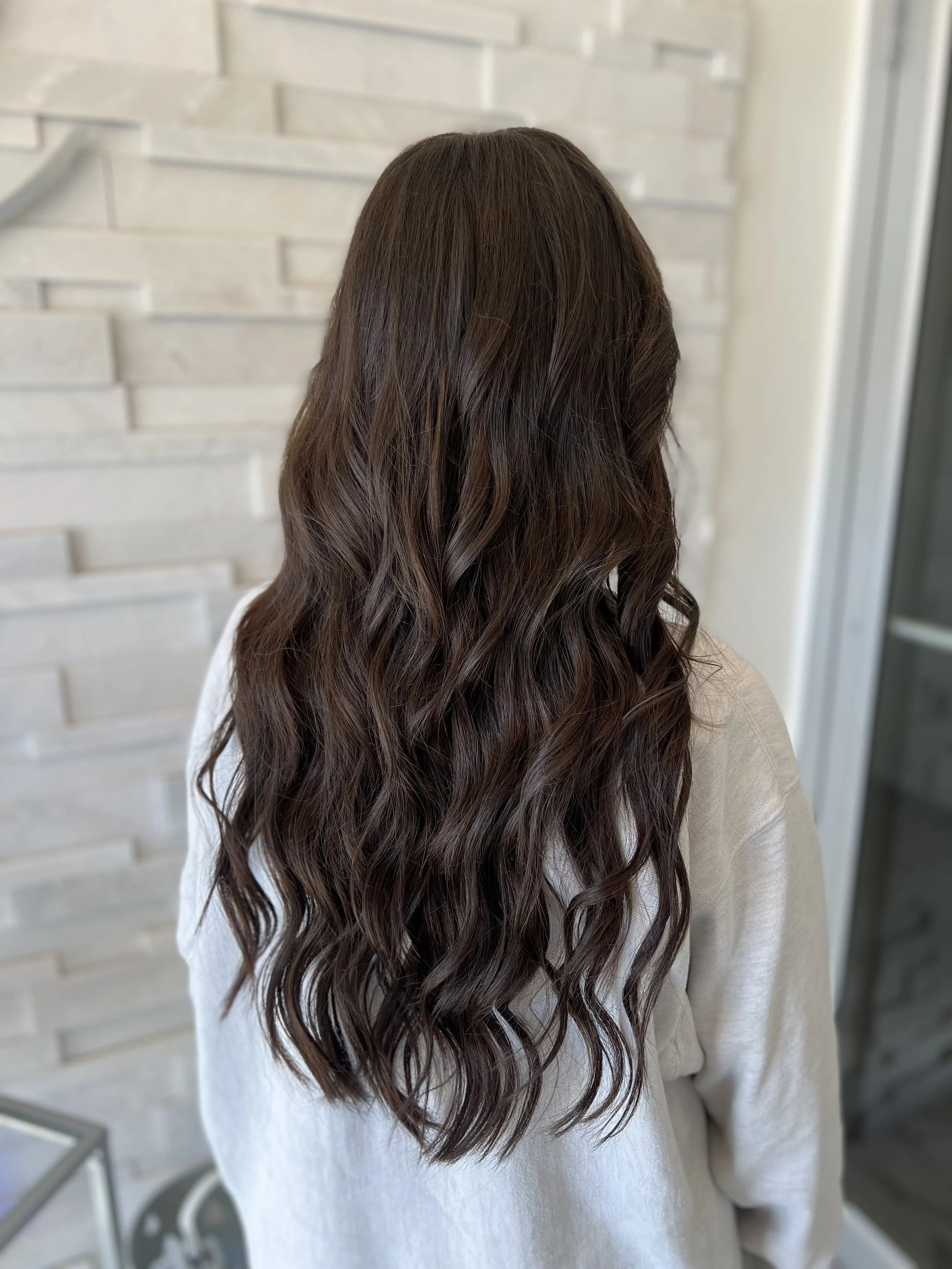 A woman with long, wavy brown hair wearing a white sweater standing against a white brick wall.