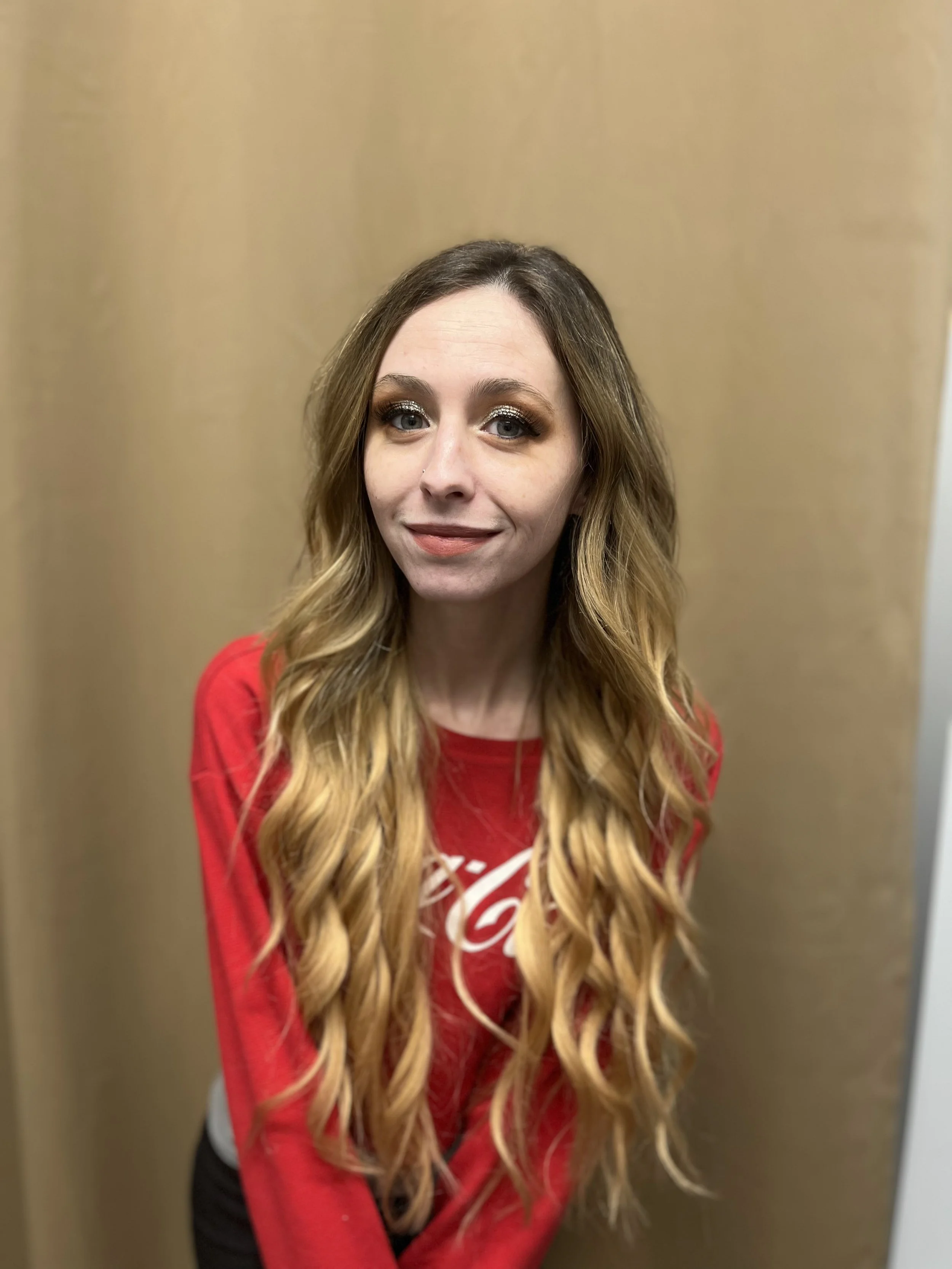 A woman with long, wavy blonde hair wearing a red shirt with a white logo, standing against a plain beige wall.