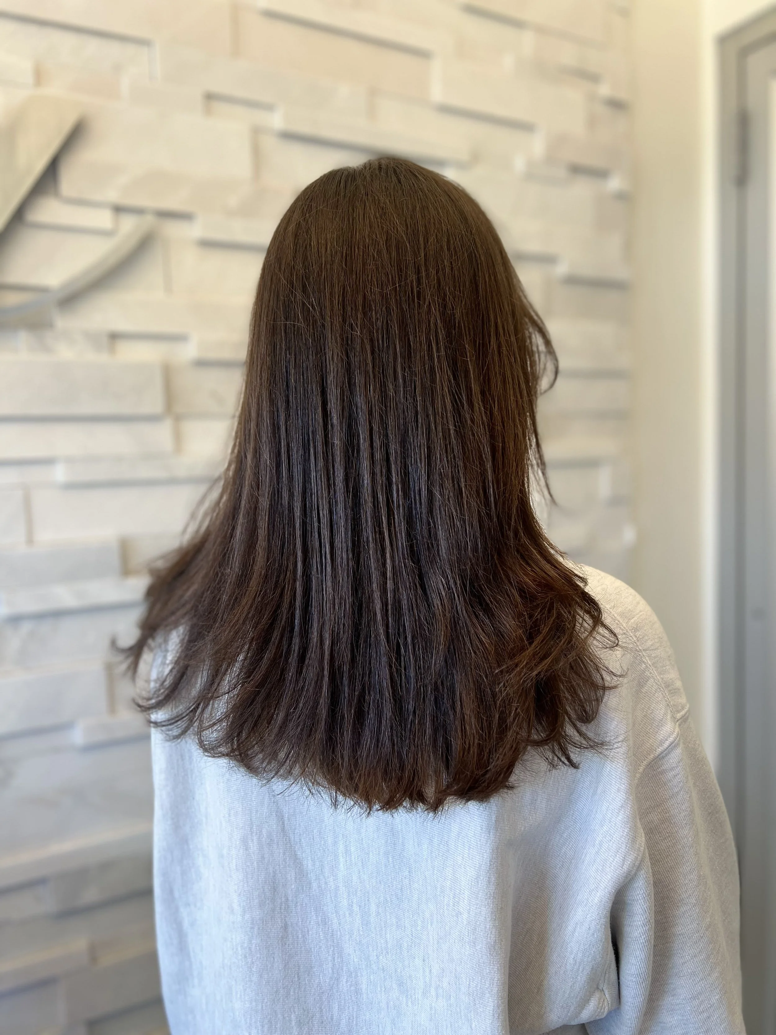 Back view of a woman with long, wavy brown hair wearing a beige sweater, standing in front of a textured white wall.