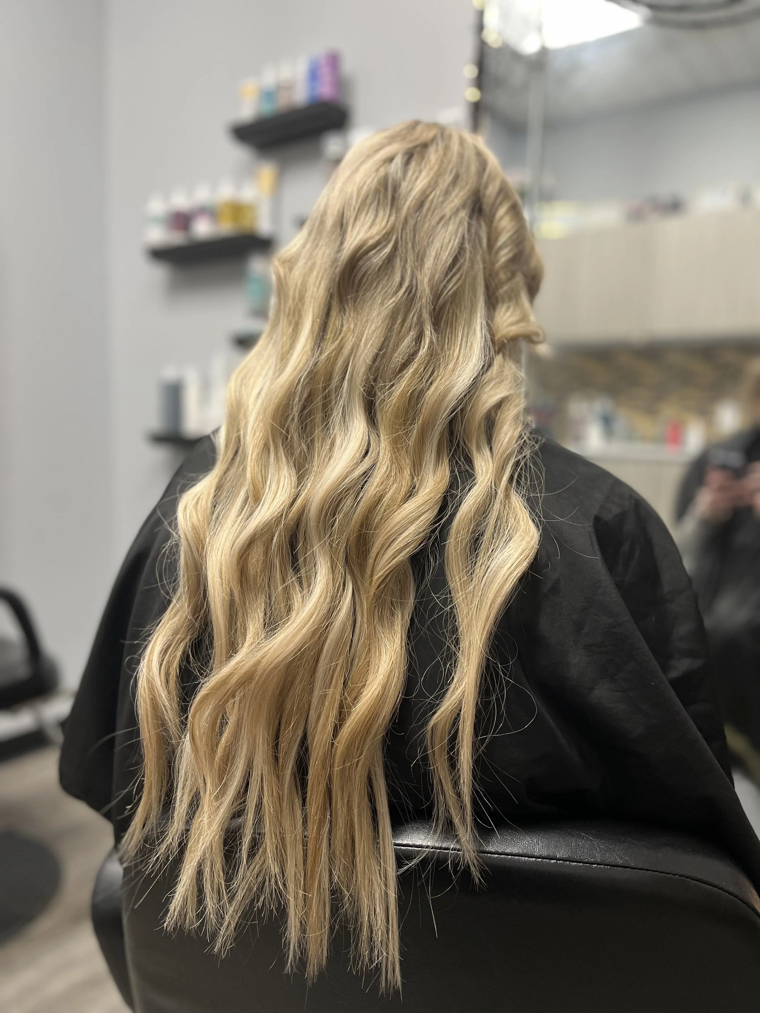 Long wavy blonde hair on a person sitting in a salon chair with shelves of hair products in the background.