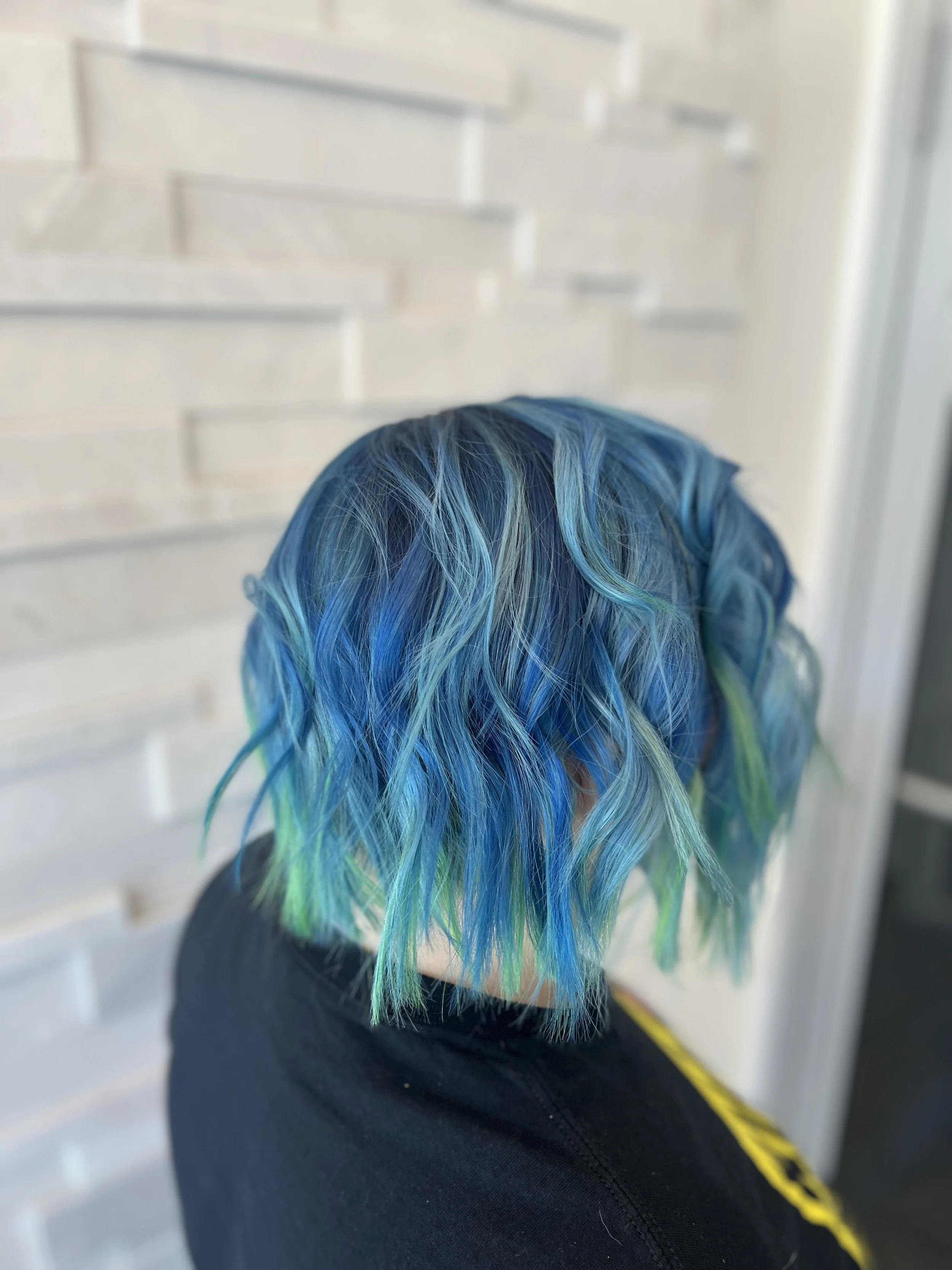 Person with short blue and green hair standing in front of a textured white wall.