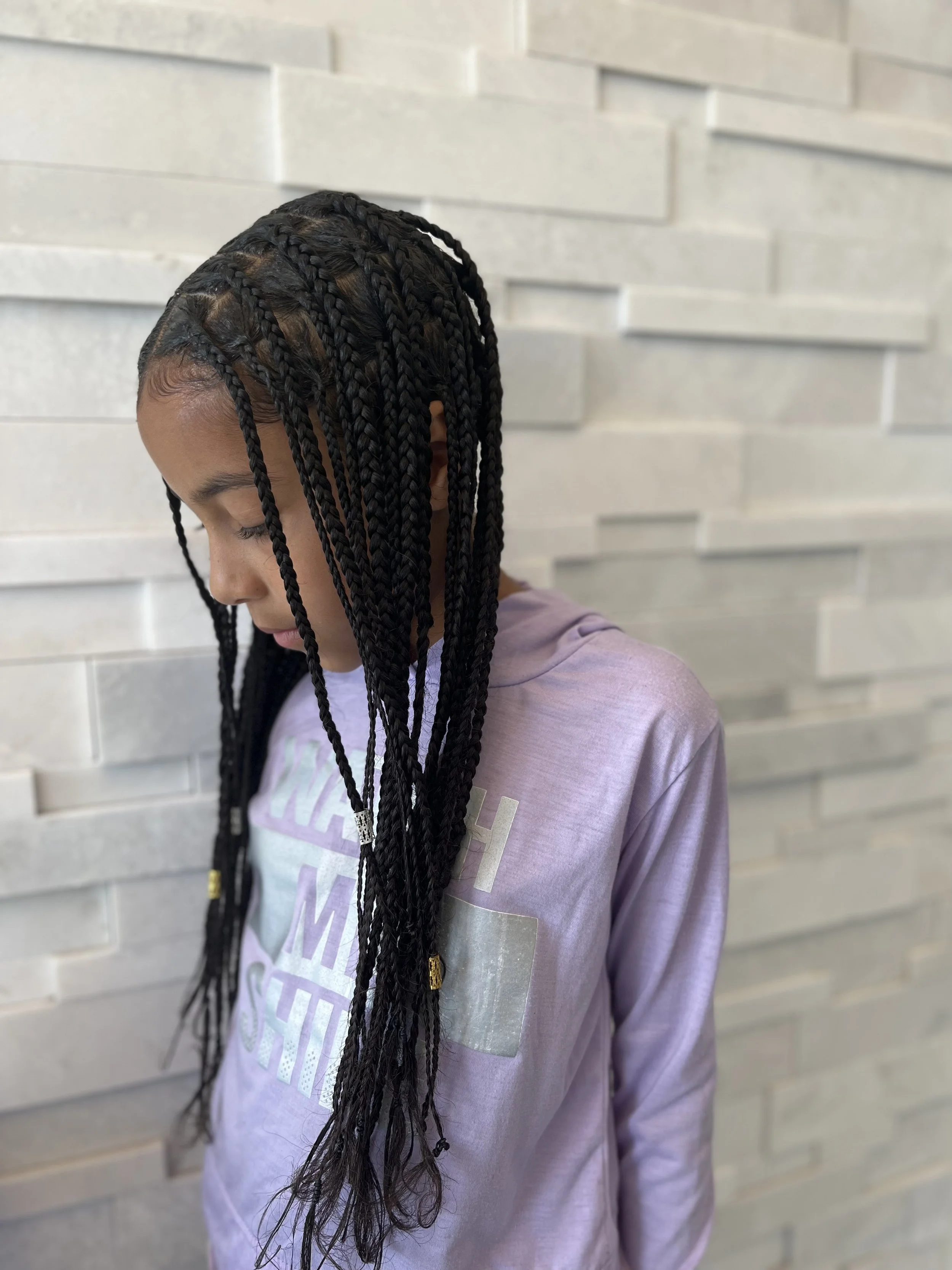 Child with long braided hair and hair accessories, wearing a light purple top, standing against a textured white wall.
